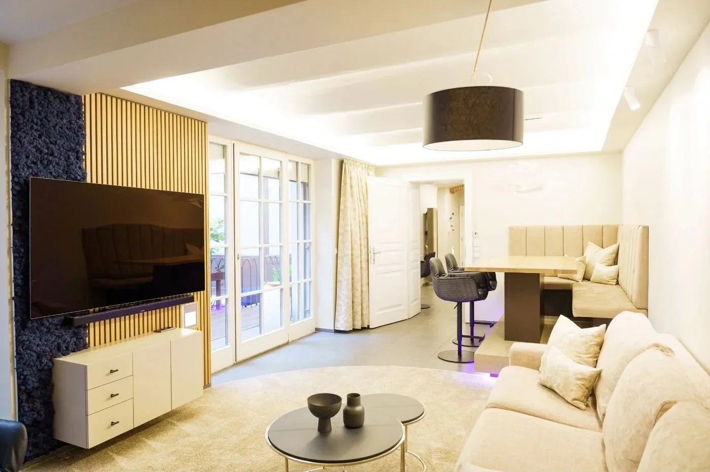 Heller Wohn- und Essbereich mit Sofa, Essnische, TV-Wand mit Holzlamellen und indirekter Deckenbeleuchtung