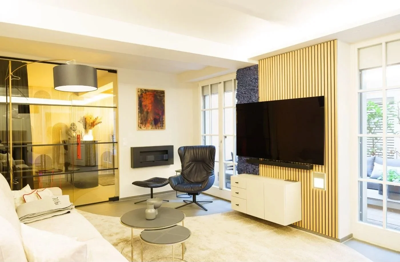 Modernes Wohnzimmer mit Luxus-Vibe: TV-Wand mit Holzlamellen und Musikbox ohne sichtbare Kabel, blaue Mooswand, Glaswand mit warmem Licht, helle Sofa-Lounge und Design-Sessel - stilvolle Renovierung