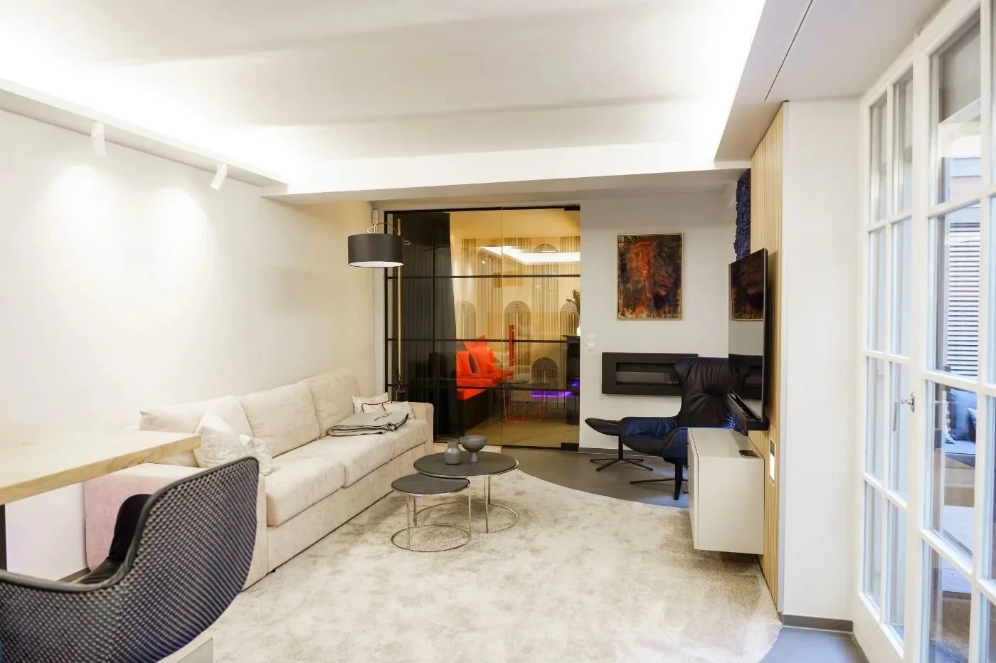 Helles, modern renoviertes Wohnzimmer mit Luxus-Vibe: große Sofa-Lounge, runder Couchtisch, Designer-Leselampe, edle Hausbar mit Glastür und roten Möbeln, Ambiente-Licht, blaues Islandmoos, Akustik-Holzpaneele, offener Essbereich mit maßangefertigten