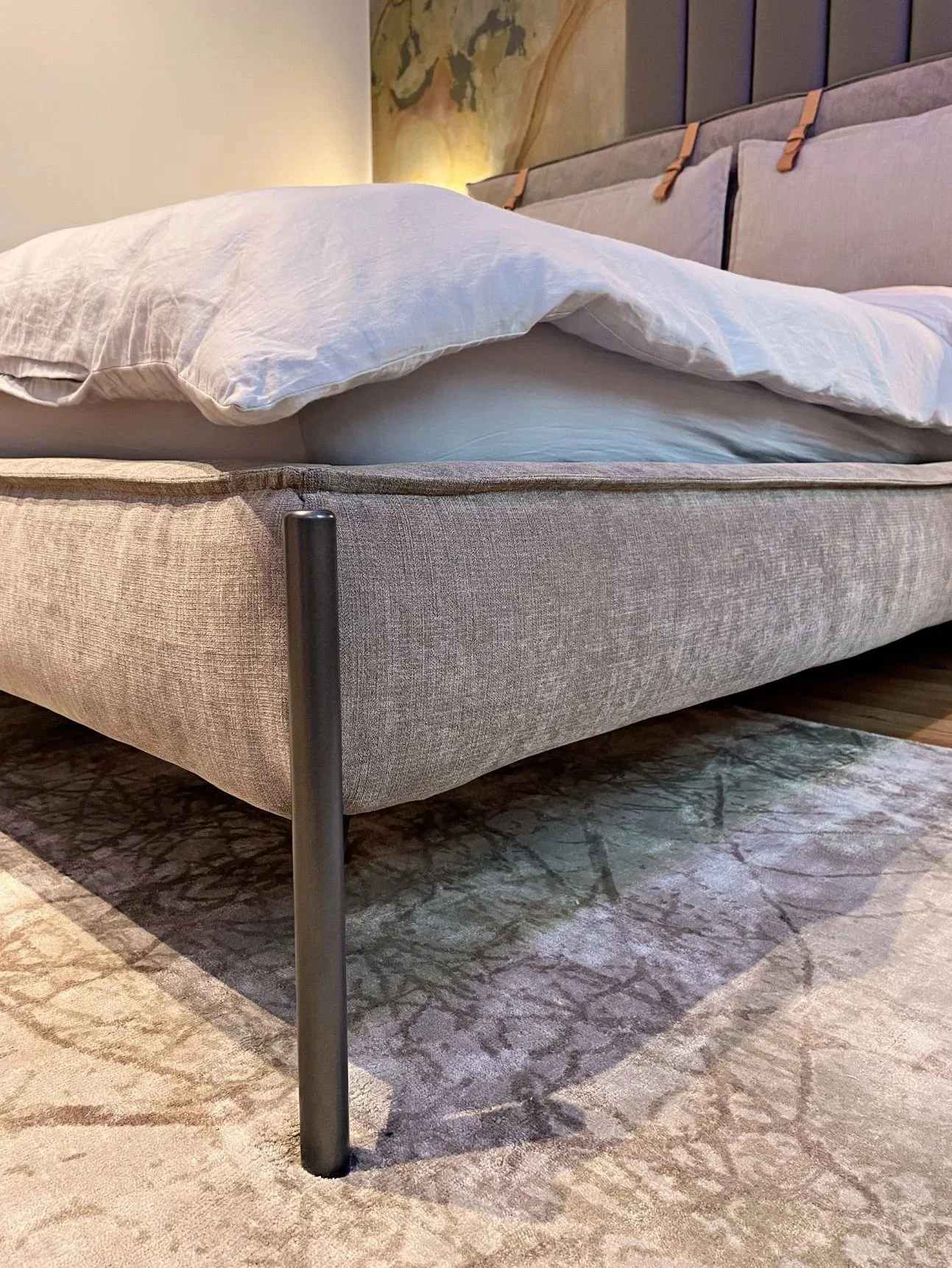 Bett mit weißer Bettwäsche und Kissen, graue Bettumrandung, Holz- und Metallbeine des Bettes, Teppichboden mit Muster im Schlafzimmer.