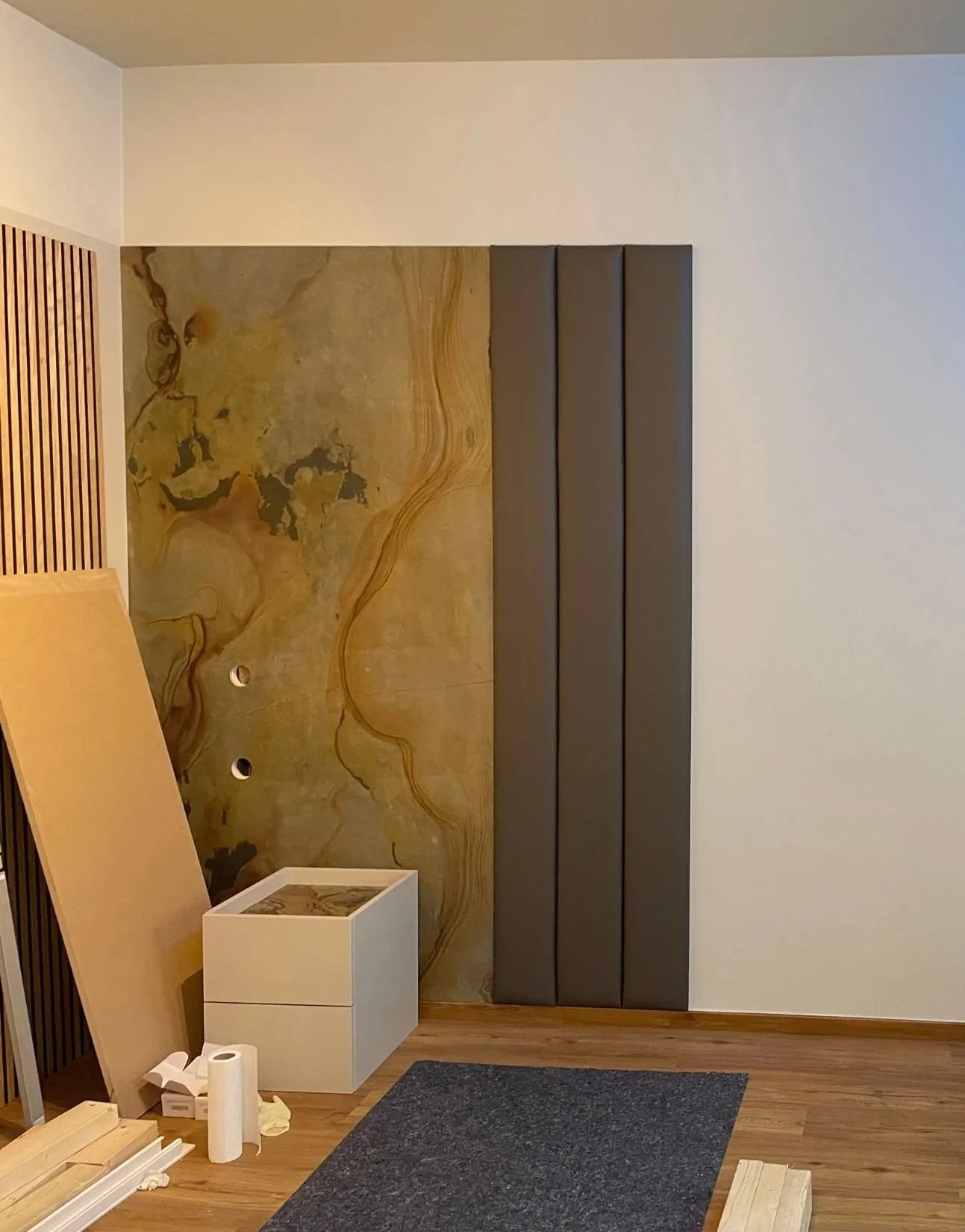 Schlafzimmer in Renovierung: Akzentwand mit Holzlamellen und Natursteinoptik im Aufbau, neue Bodenfläche und vorbereitete Möbelmontage - moderne Renovierung der Paddy Artist Design GmbH
