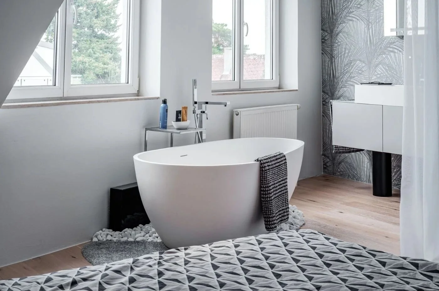 Modernes Schlafzimmerbad mit freistehender Badewanne, hellen Fenstern, Holzfußboden und klarer Gestaltung in Weiß und Grau – stilvolle Badarchitektur mit wohnlicher Atmosphäre, offenen Sichtachsen und hochwertigem Spa-Charakter
