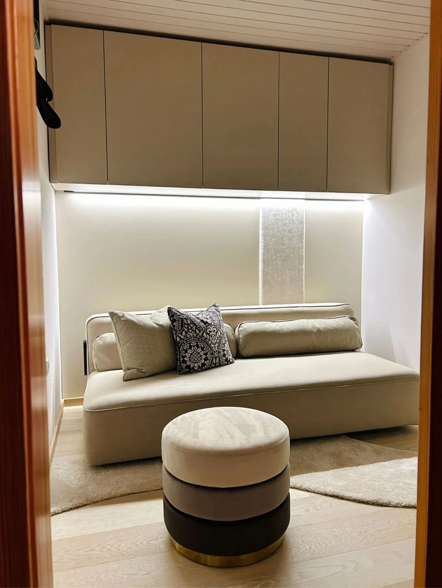 Beleuchtete Wohnnische mit hellem Sofa, Kissen und rundem Polsterhocker.