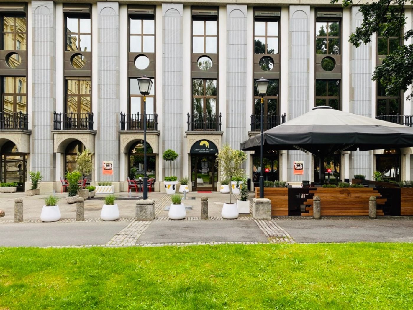 Großzügige Ansicht einer markanten Restaurantfassade mit Arkaden, Stadtterrasse, bepflanzten Elementen und überdachtem Gastgarten in Maribor - stilvoll gestalteter Außenbereich mit urbanem Flair, grünem Vorplatz und einladender Atmosphäre