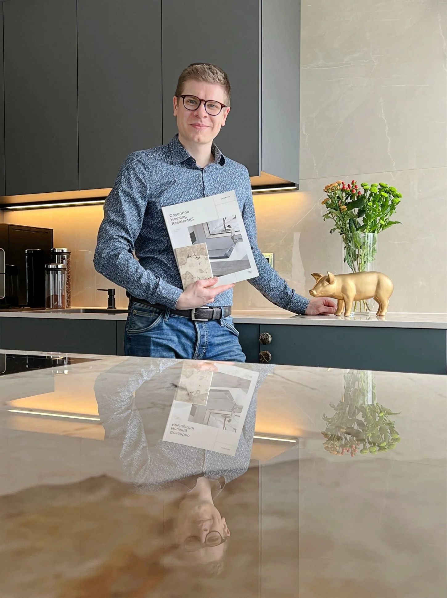 Berater steht in einer modernen, mattschwarzen Designküche mit warmem Licht, hält eine Planungsmappe in der Hand und zeigt eine maßgefertigte Küchenlösung mit Stein-Arbeitsplatte - Küchenplanung, Küchenkauf und Beratung in Wien und Umgebung