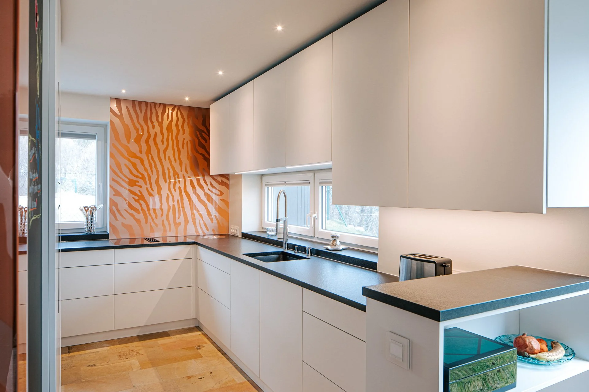 Moderne Einbauküche mit weißen, grifflosen Fronten, schwarzer Arbeitsplatte und auffälliger, orange gemusterter Küchenrückwand - hochwertiges Küchen- und Innenraumdesign