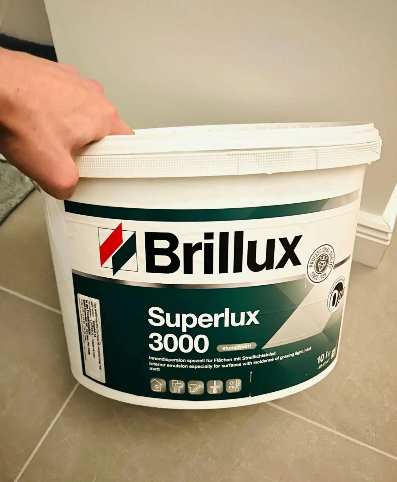 Ein Eimer Brillux Superlux 3000 Wandfarbe mit grünem Etikett und Hand, die den Deckel anhebt, steht auf einer Fliese. Im Hintergrund eine weiße Wand.