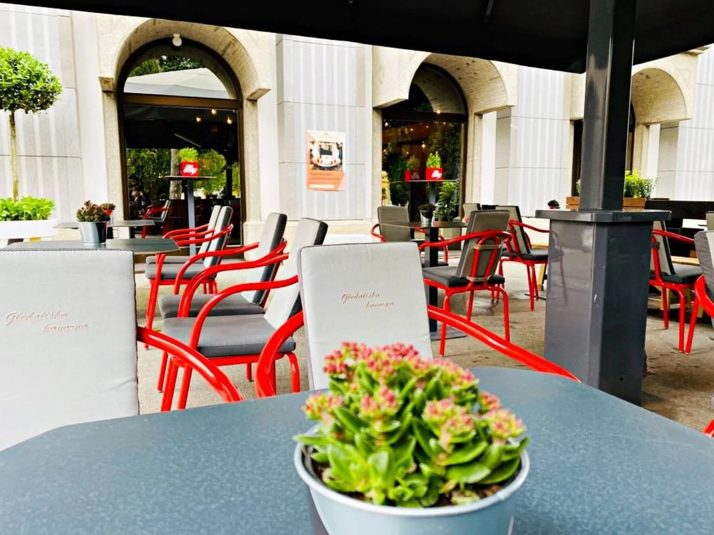 Moderner Gastgarten mit roten Stühlen, schwarzen Tischen und bepflanzter Tischdekoration vor Arkadenfassade in Maribor - stilvoller Restaurant-Außenbereich mit geschützter Terrasse und einladender Atmosphäre