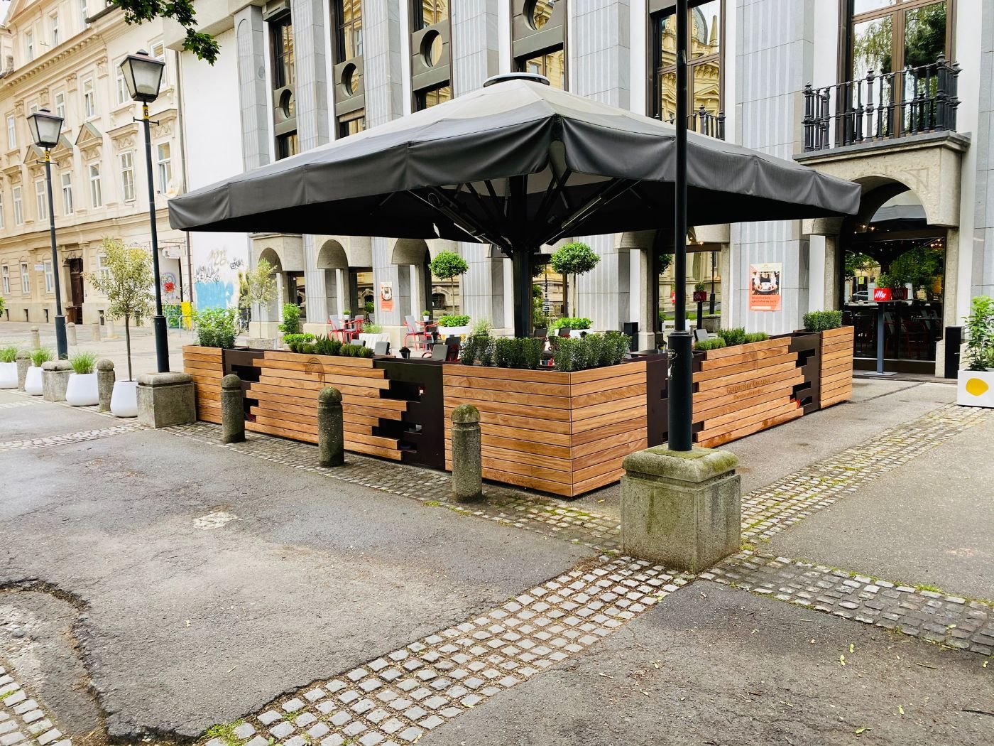 Moderner Gastgarten mit Holzverkleidung, bepflanzten Elementen und großem schwarzem Sonnenschirm in urbaner Altstadtlage in Maribor - stilvoll gestalteter Restaurant-Außenbereich mit geschützter Stadtterrasse und einladender Atmosphäre