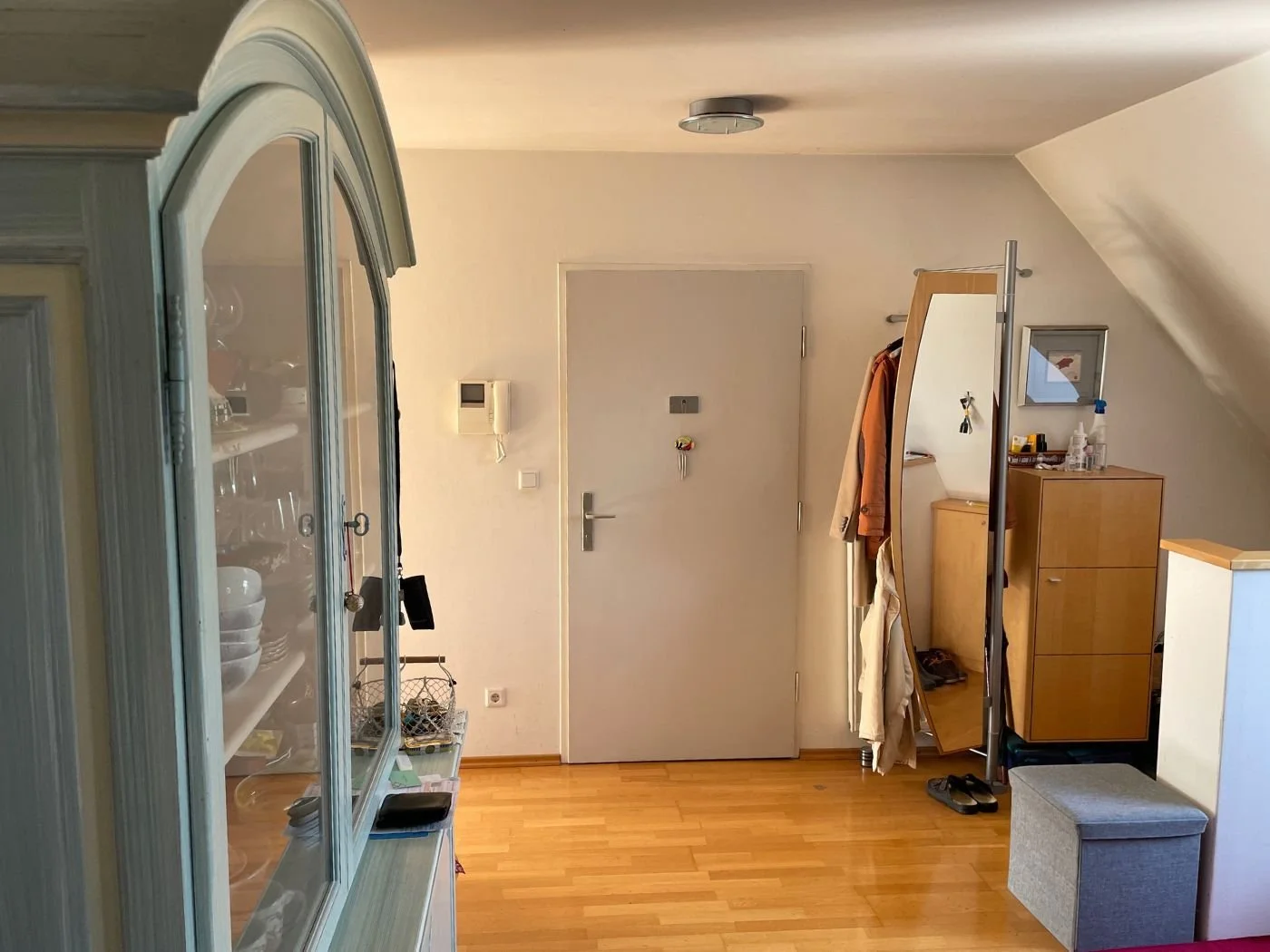 Bild eines Flurs im Bestandszustand mit Vitrine, offener Garderobenlösung, Sitzhocker und einfacher Eingangssituation unter Dachschräge – Ausgangssituation vor der Neugestaltung mit mehr Stauraum, klarerer Raumstruktur und moderner Innenarchitektur