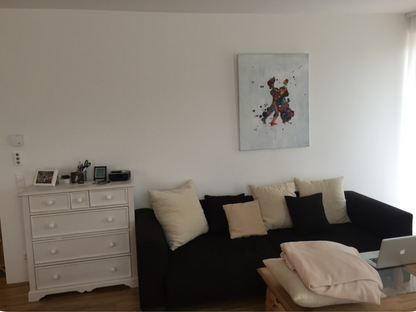 Vorher-Bild eines Wohnzimmers mit weißer Wand, dunklem Sofa und schlichter Einrichtung vor der gestalterischen Neuausrichtung