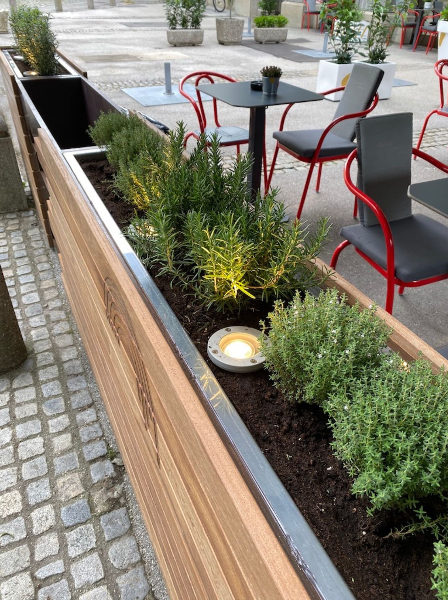 Langer Pflanzenkasten mit Holzverkleidung, Kräuterbepflanzung und integrierter Beleuchtung im modernen Gastgarten vor roten Stühlen und schwarzen Tischen in Maribor - stilvoll gestalteter Restaurant-Außenbereich mit natürlicher Begrünung und urbanem 