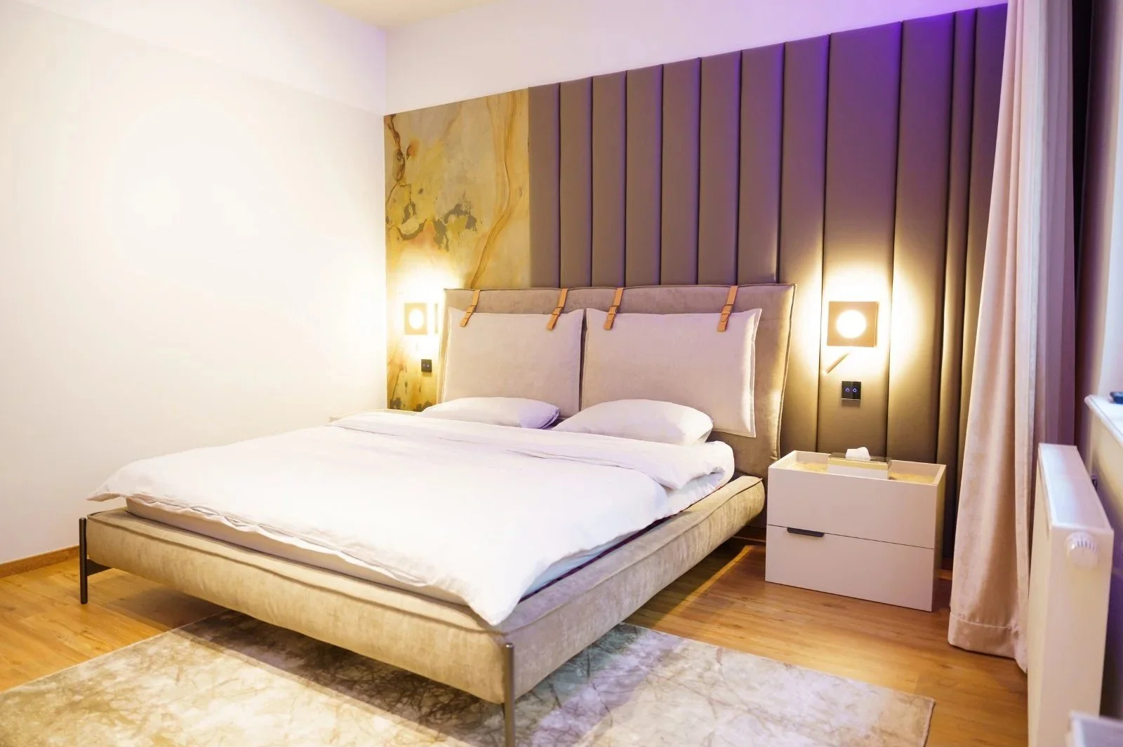 Schlafzimmer mit beigefarbenem Polsterbett, violetter Akzentwand und indirekter Beleuchtung