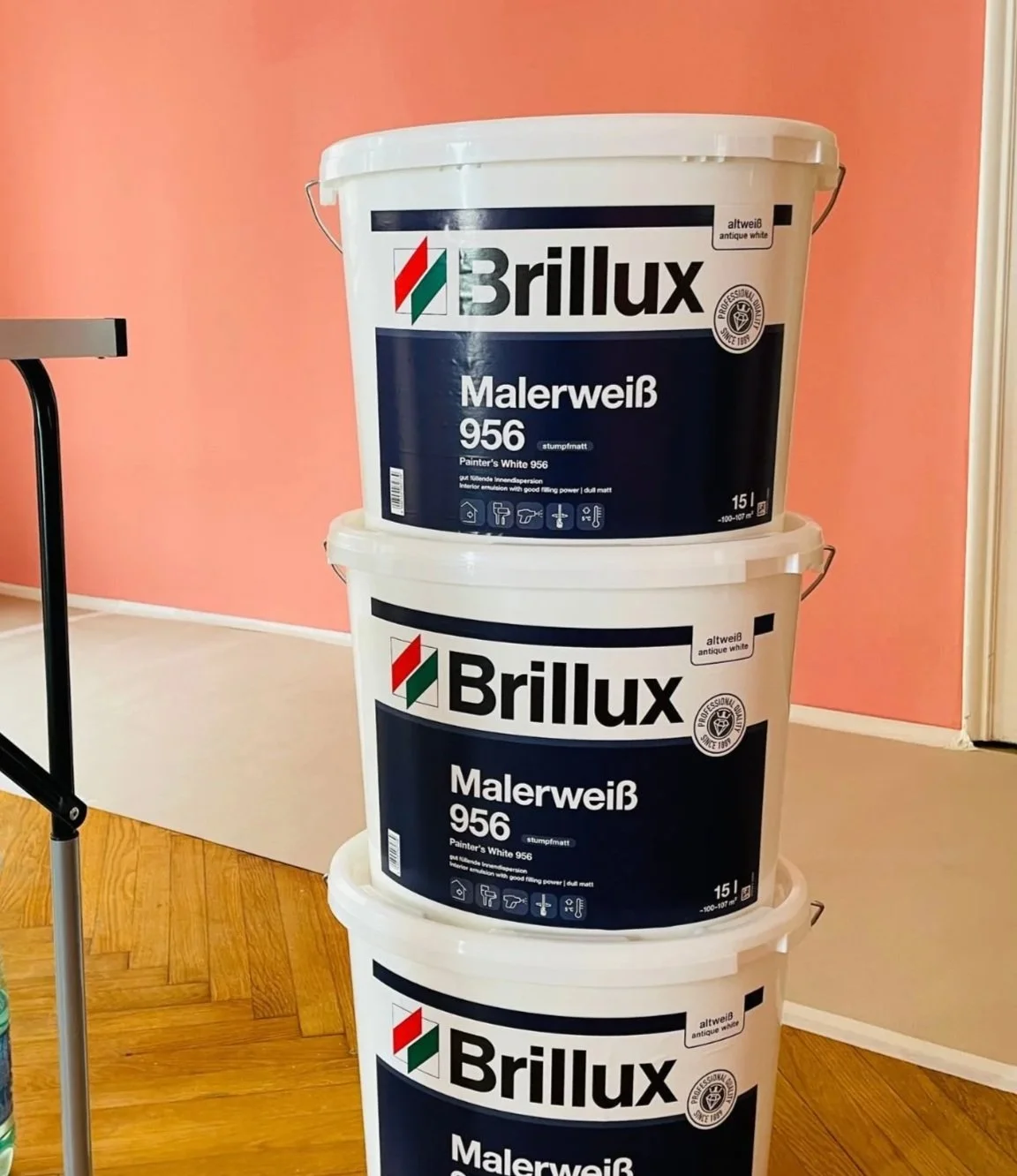 Drei Eimer mit weißer Farbe von der Marke Brillux, gestapelt in einem Raum mit rotem und beigem Wand, Holzboden und einem Tisch an der Seite.