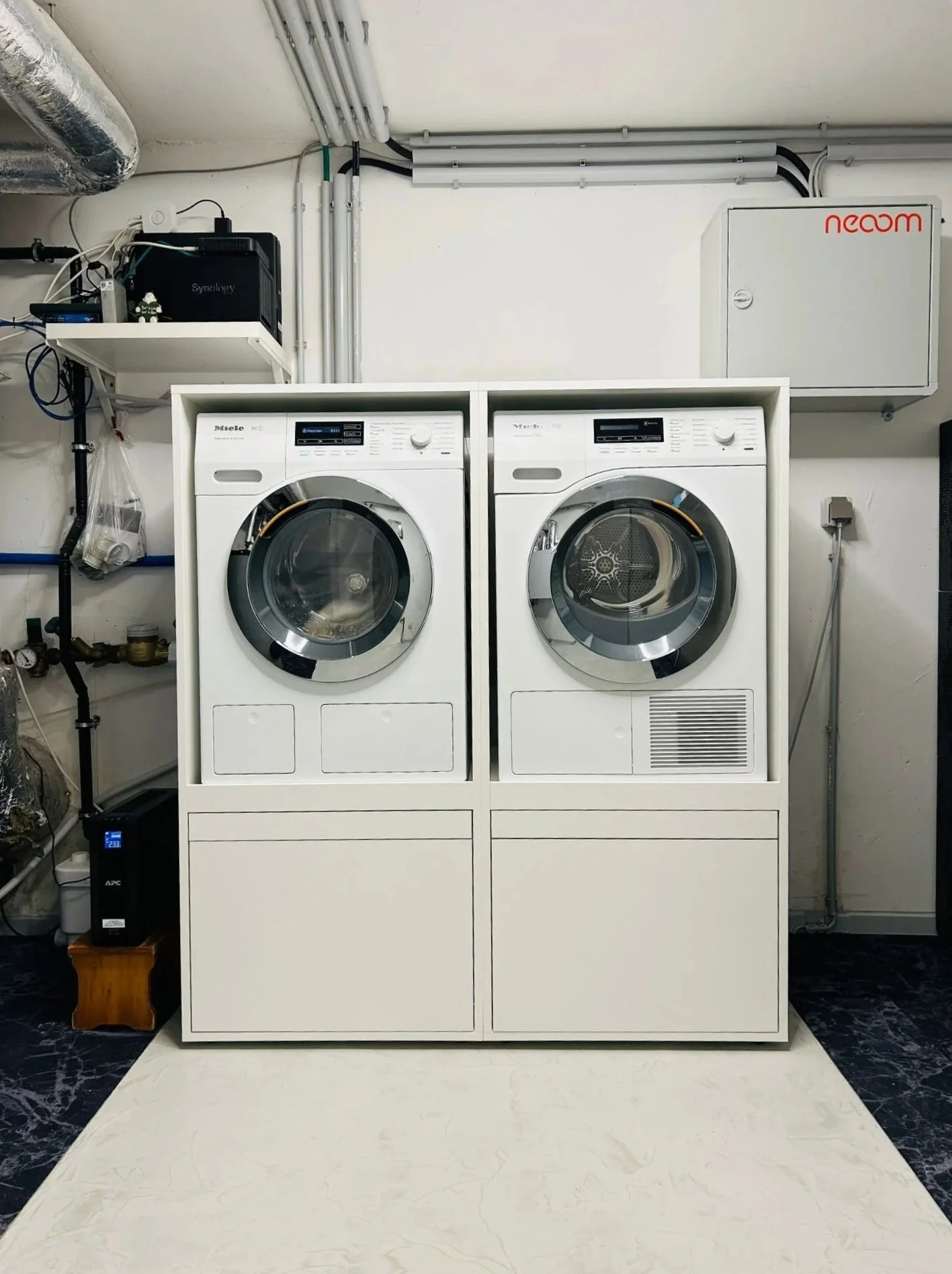 Waschmaschine und Trockner in einer weißen Möbelbox mit schwarzem Boden, in einer Waschküche, im Hintergrund ein grauer Kabelkanal und ein grauer Sicherungskasten