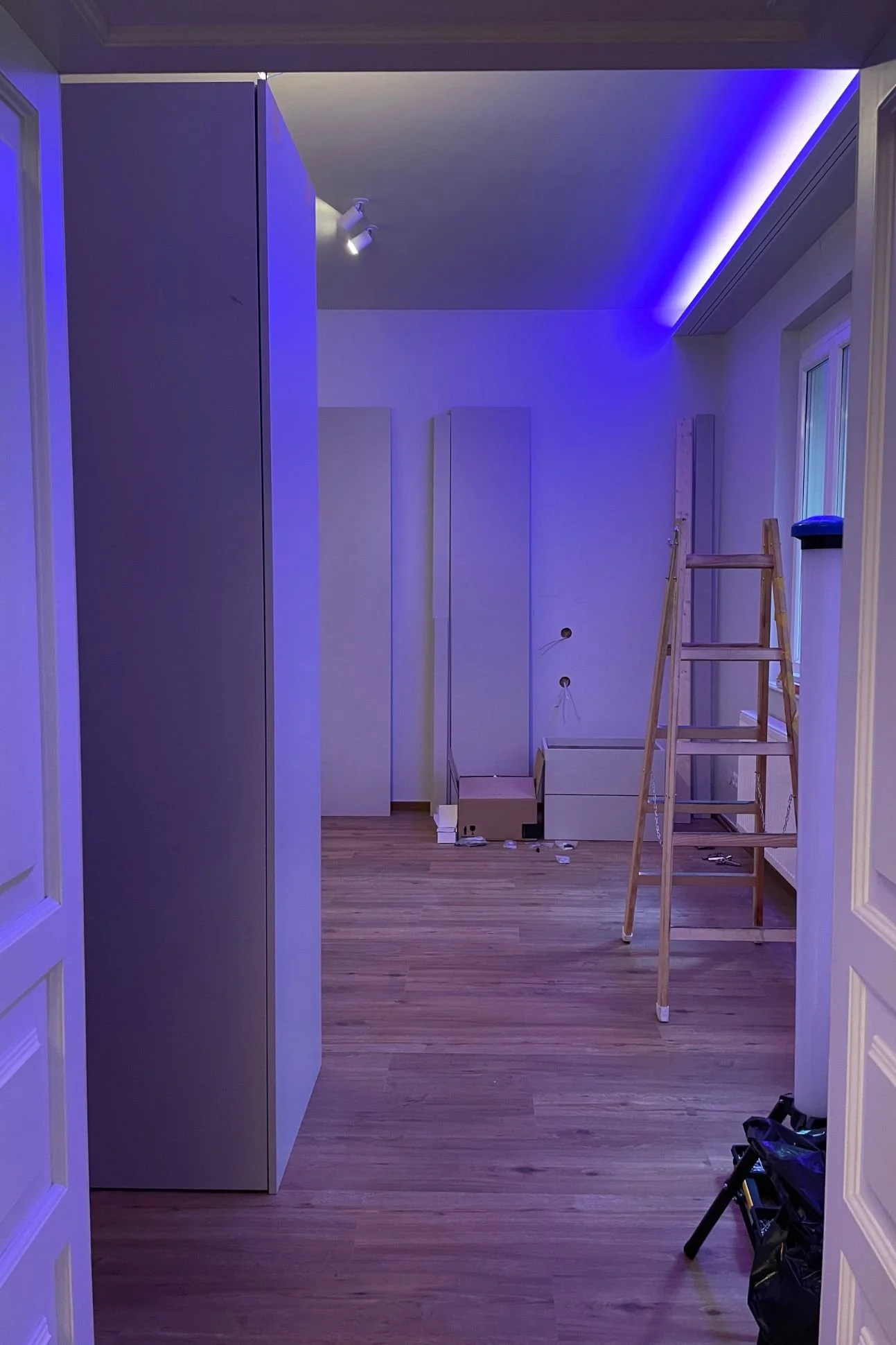 Schlafzimmer während der Renovierung im Umbau: Boden verlegt, Möbelmontage und Leiter im Raum, indirekte LED-Beleuchtung mit blauem Stimmungslicht - moderne Renovierung der Paddy Artist Design GmbH