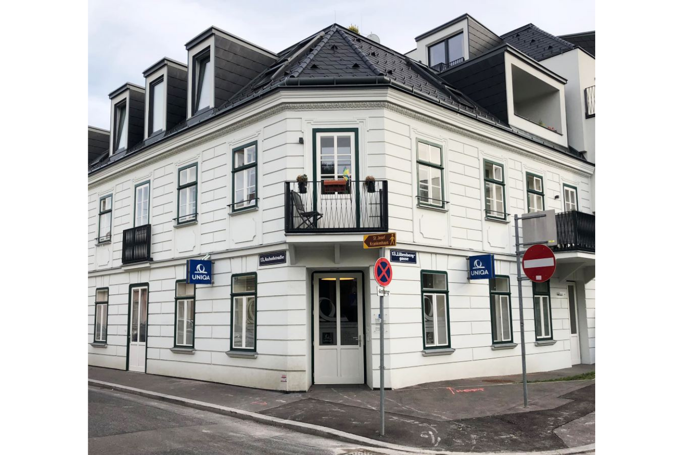 Mehrstöckiges weißes Gebäude mit schwarzen Fensterschienen und einem kleinen Balkon. Verkehrsschilder an der Ecke, darunter Einbahnstraßenschilder und Parkverbot. Straßennamen auf blauen Schildern. Eine Ecke der Straße mit Gehweg und Autos auf der Straße.