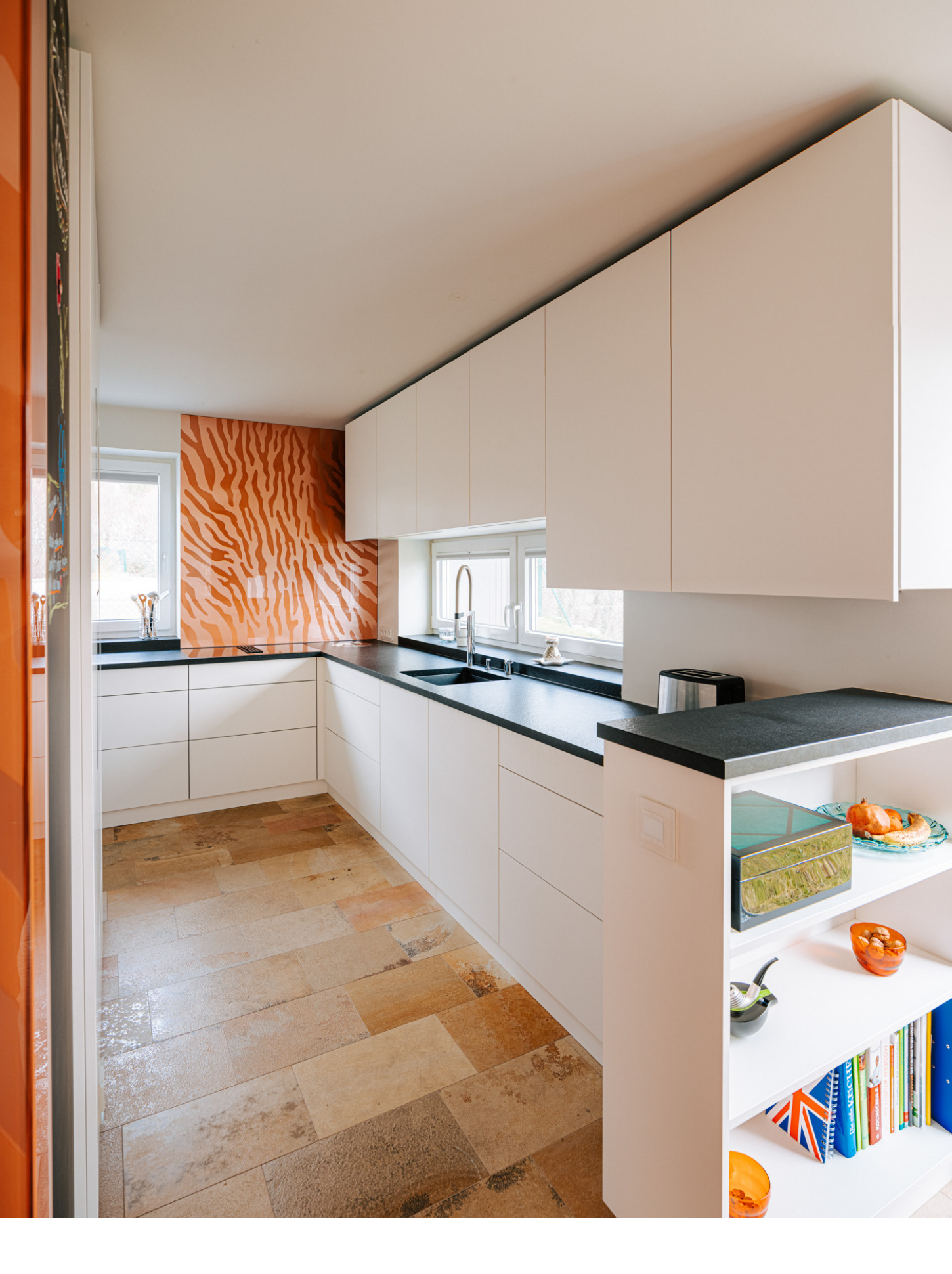Weitwinkelaufnahme einer modernen, L-förmigen Küche mit weißen Fronten und schwarzer Arbeitsplatte, Spüle am Fenster und auffälliger, orange gemusterter Rückwand als Highlight - Küchenplanung und Küchenrenovierung vom Meisterbetrieb