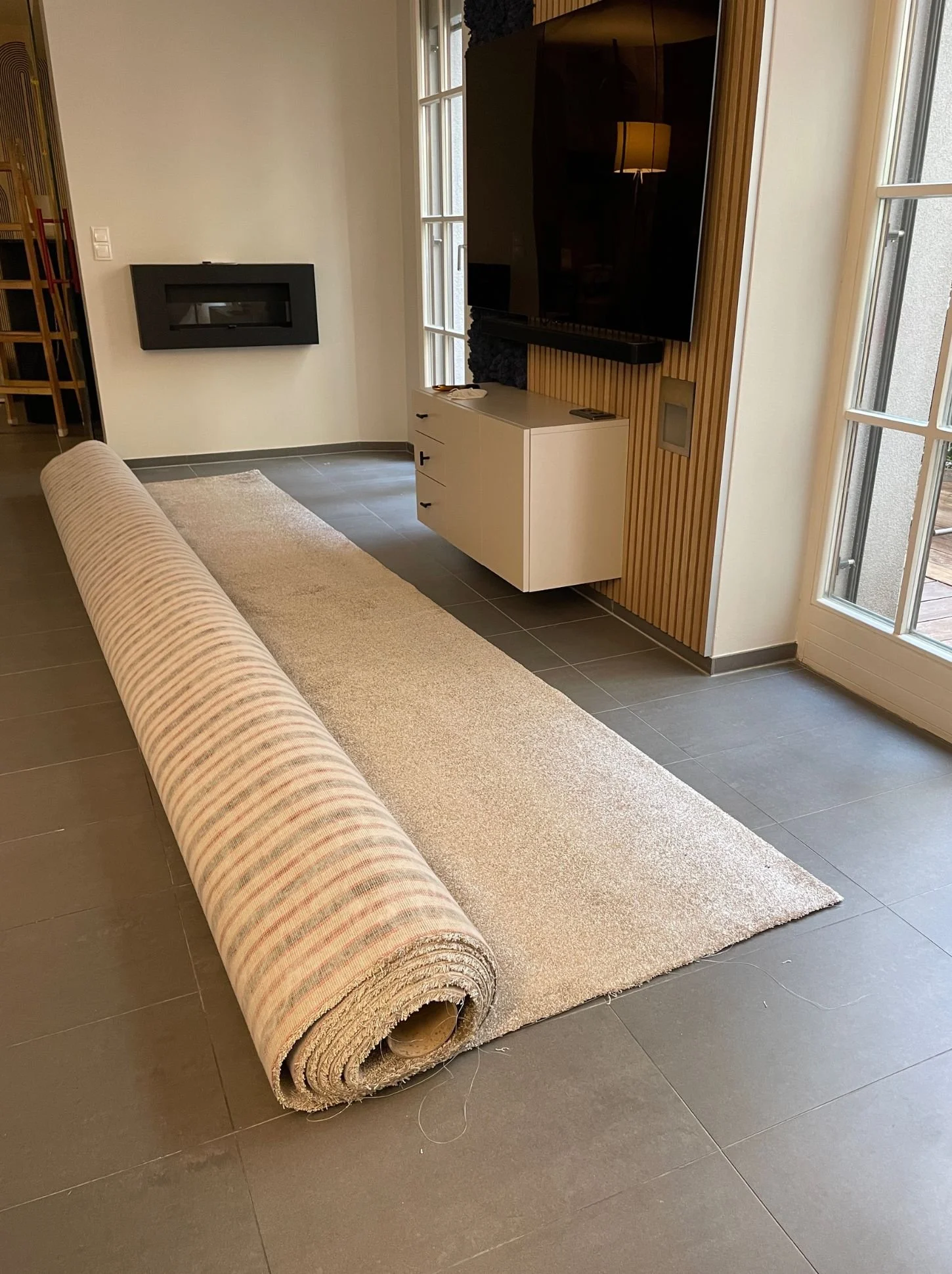 Ein eingewickelter Teppich liegt auf einem Fliesenboden in einem Wohnzimmer, mit einem weißen Sideboard, einem großen Fernseher und einer Fensterfront im Hintergrund.