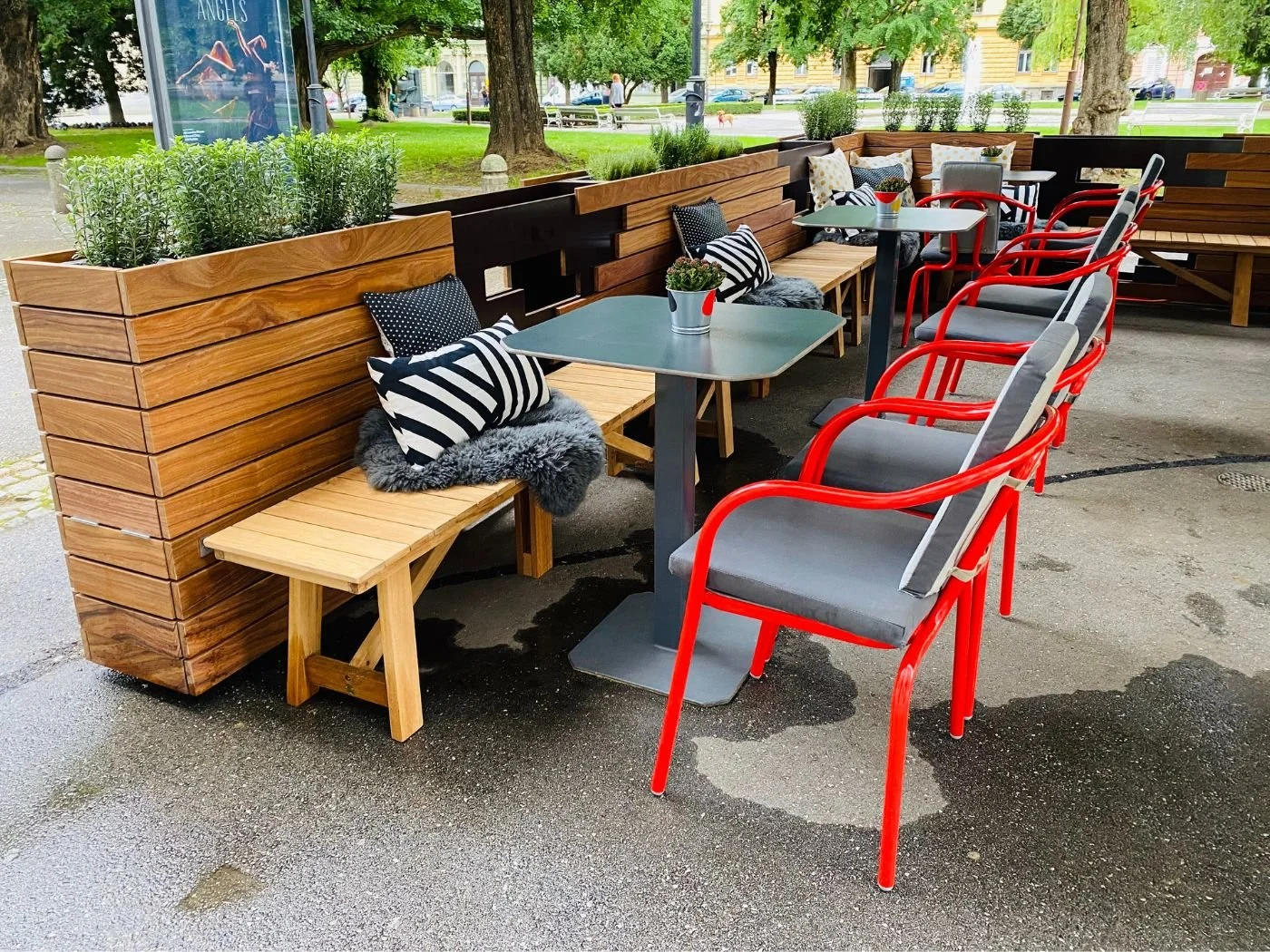 Moderner Gastgarten des Theatercafes Gledaliska Kavarna mit Holzbank, integriertem Planzenkasten, schwarzen Tischen und roten Stühlen vor einer grünen Parkkulisse in Maribor an der Drau - stilvoll gestalteter Restaurant-Außenbereich mit einladenden S