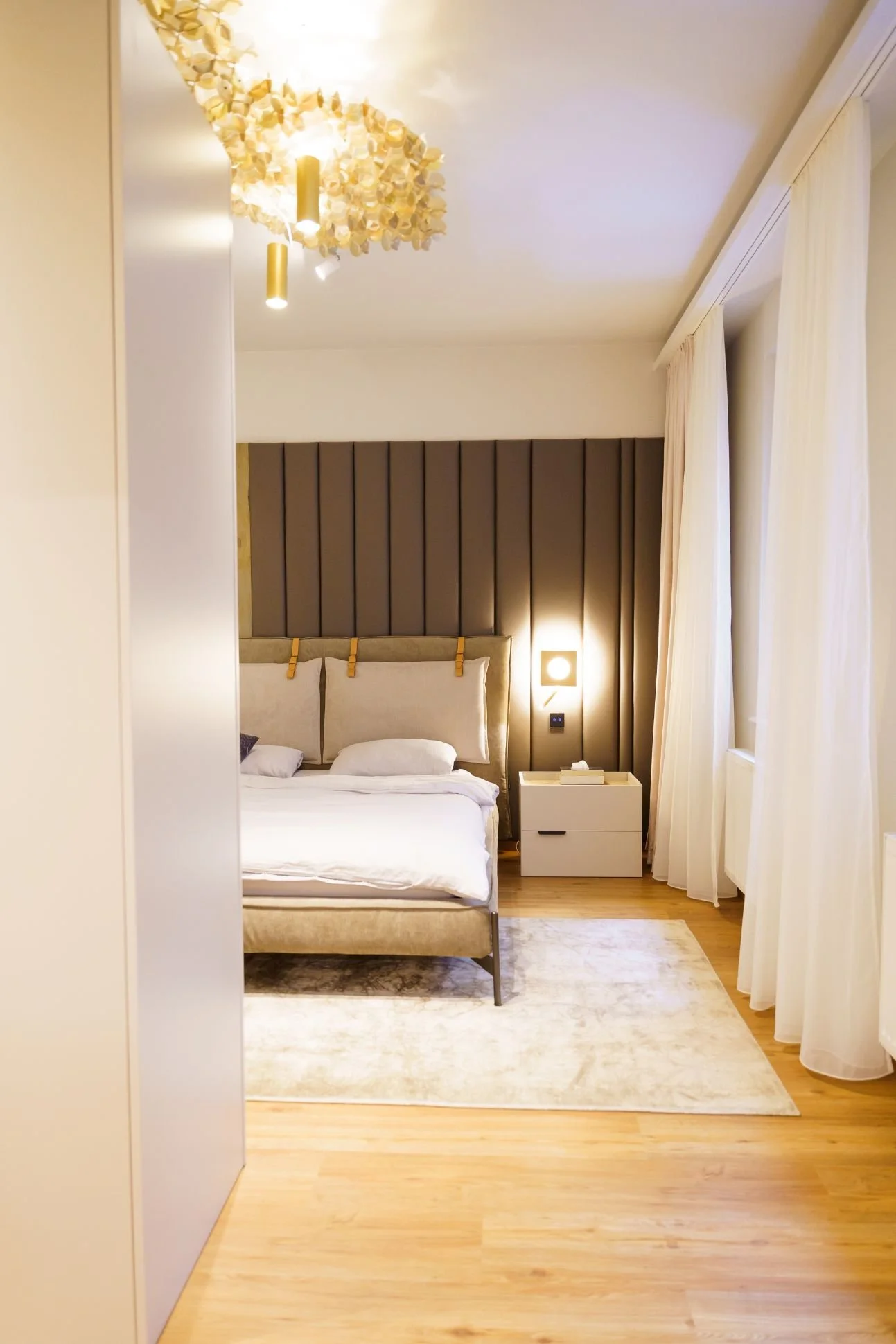 Helles Schlafzimmer mit gepolstertem Bett, Nachttisch und raumhohen Vorhängen.