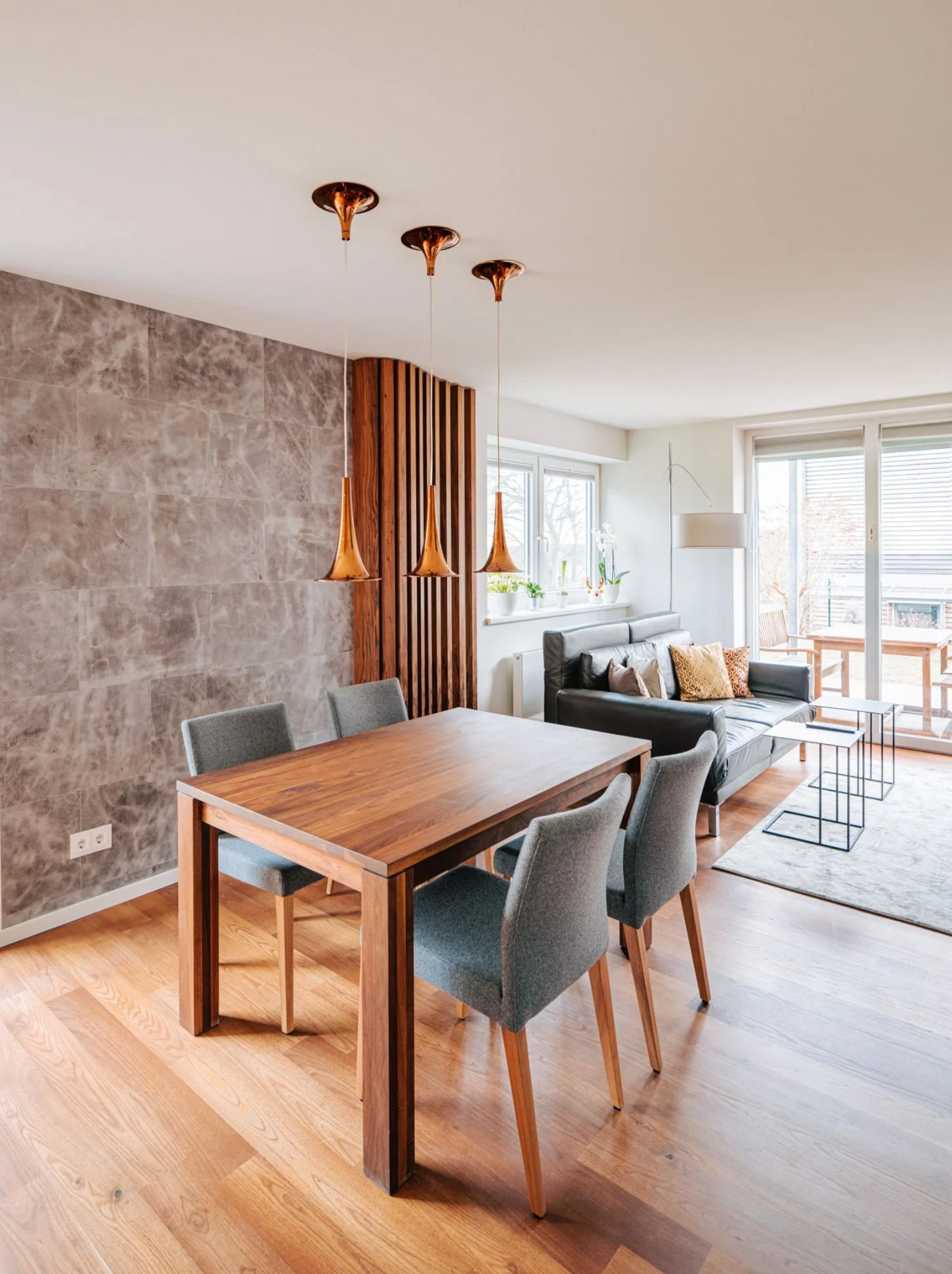 Offener Wohn- und Essbereich mit Holztisch, grauen Stühlen, dunklem Sofa, Wand in Steinoptik und Holzlamellen