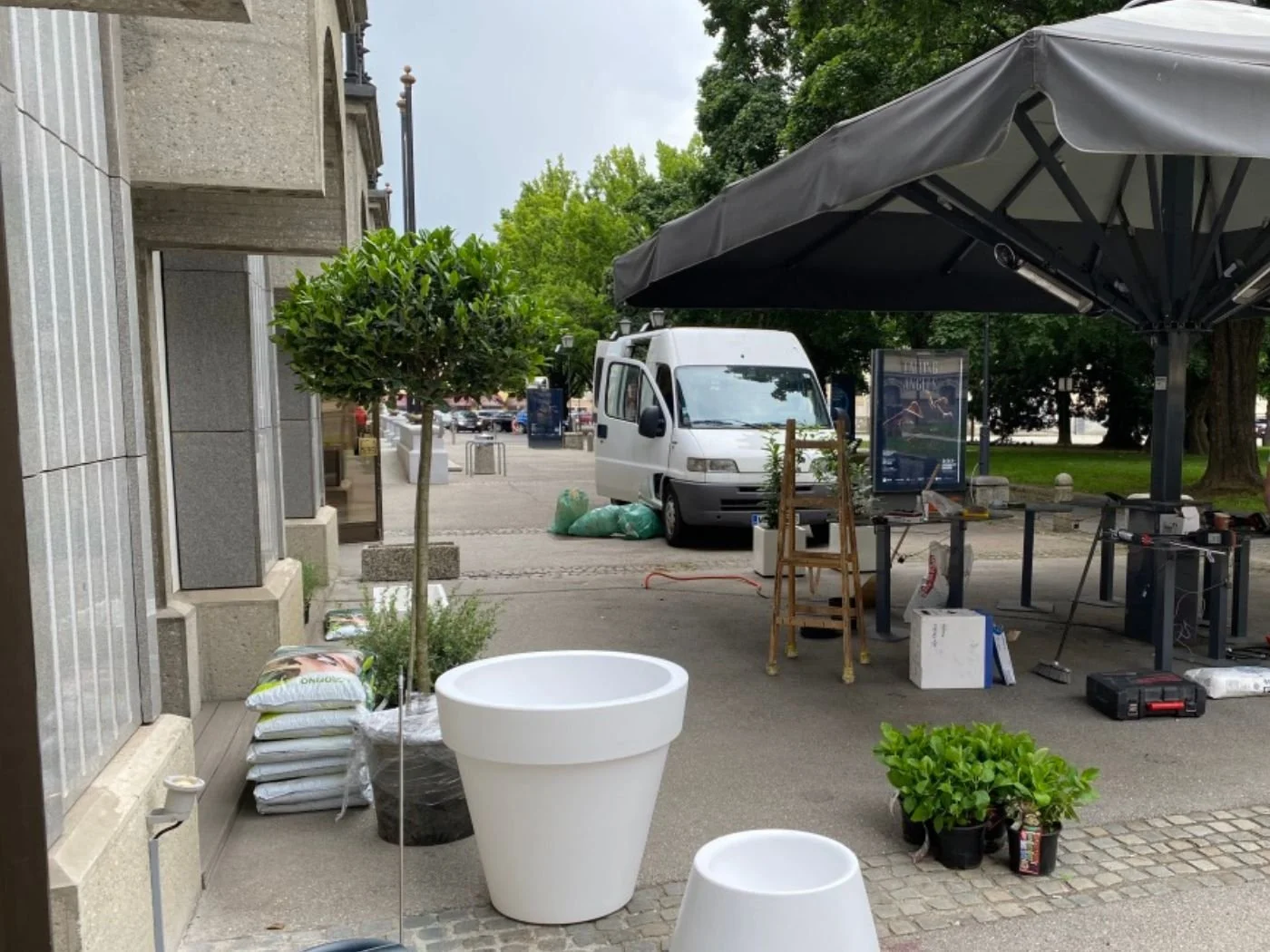 Aufbau eines modernen Gastgartens mit großen weißen Pflanzkübeln, bepflanzten Elementen und schwarzem Sonnenschirm vor einer Restaurantfassade in Maribor – hochwertig gestalteter Außenbereich mit urbaner Terrasse, natürlicher Begrünung und stilvoller
