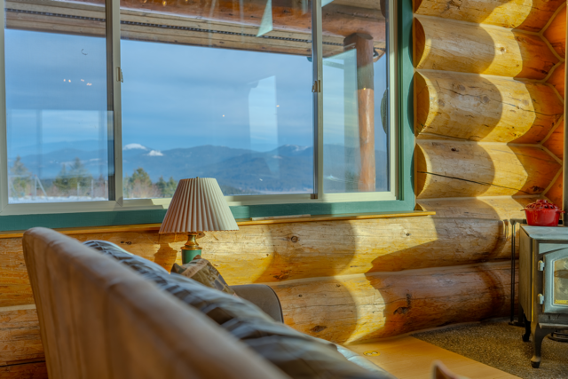 Interior of a Spokane area log cabin with a window overlooking mountains and blue sky, a lamp on a side table, and part of a sofa.