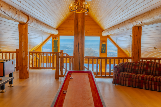 Interior of a Spokane area cozy log cabin with wooden beams, a red shuffleboard, plaid sofa, and large windows showing a mountain view.