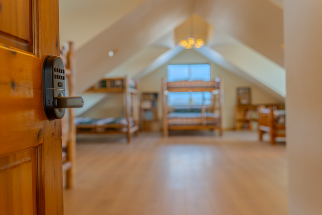View of an attic bedroom with sloped ceilings, wooden furniture including beds and bookshelves, a window with curtains, and a hanging light fixture.