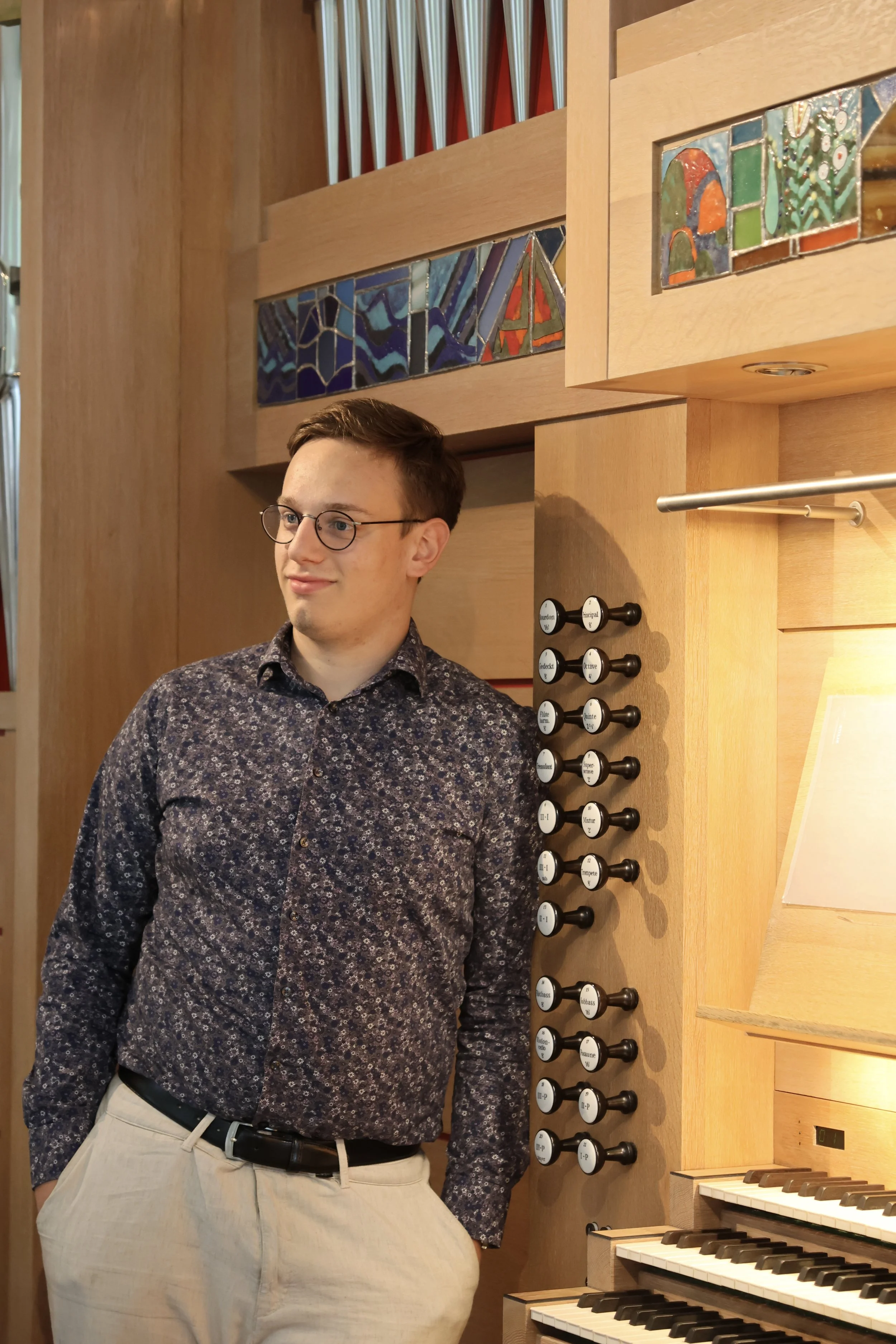 Ein junger Mann mit Brille steht in einem Raum neben einer Orgel. Er trägt ein dunkelblaues Hemd mit Blumenmuster und hellbeige Hosen.