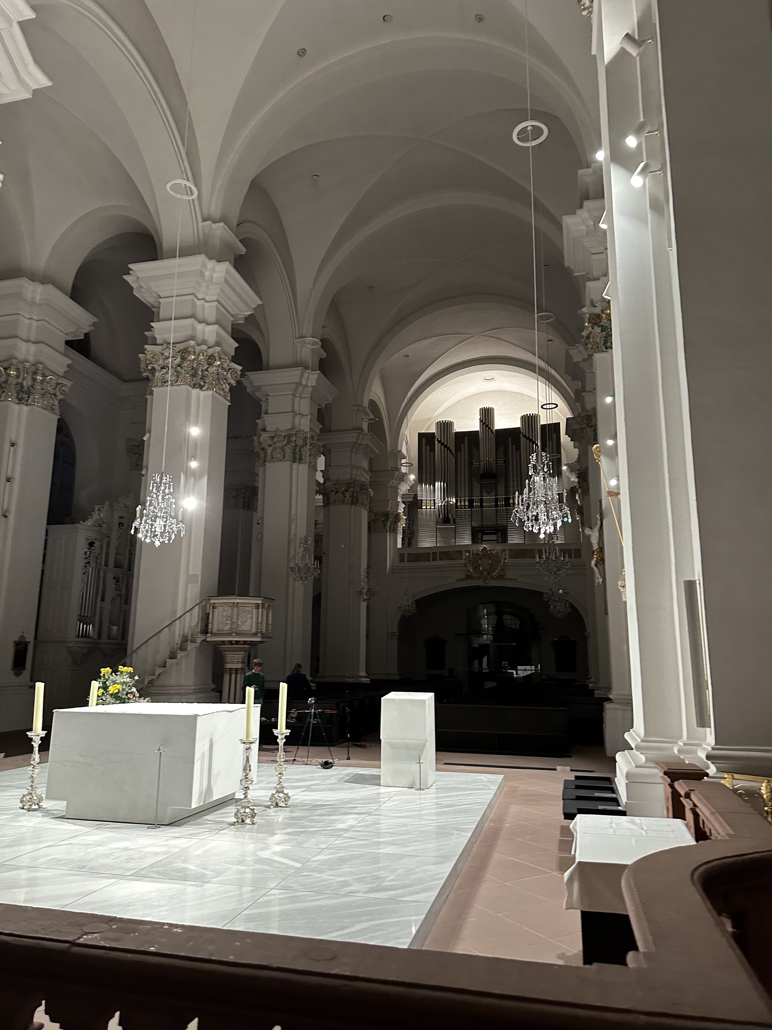 Innenansicht einer gotischen Kirche mit hohen weißen Säulen, einer großen Orgel im oberen Bereich, Kronleuchtern und einem Altar mit Kerzen und Blumen im Vordergrund.
