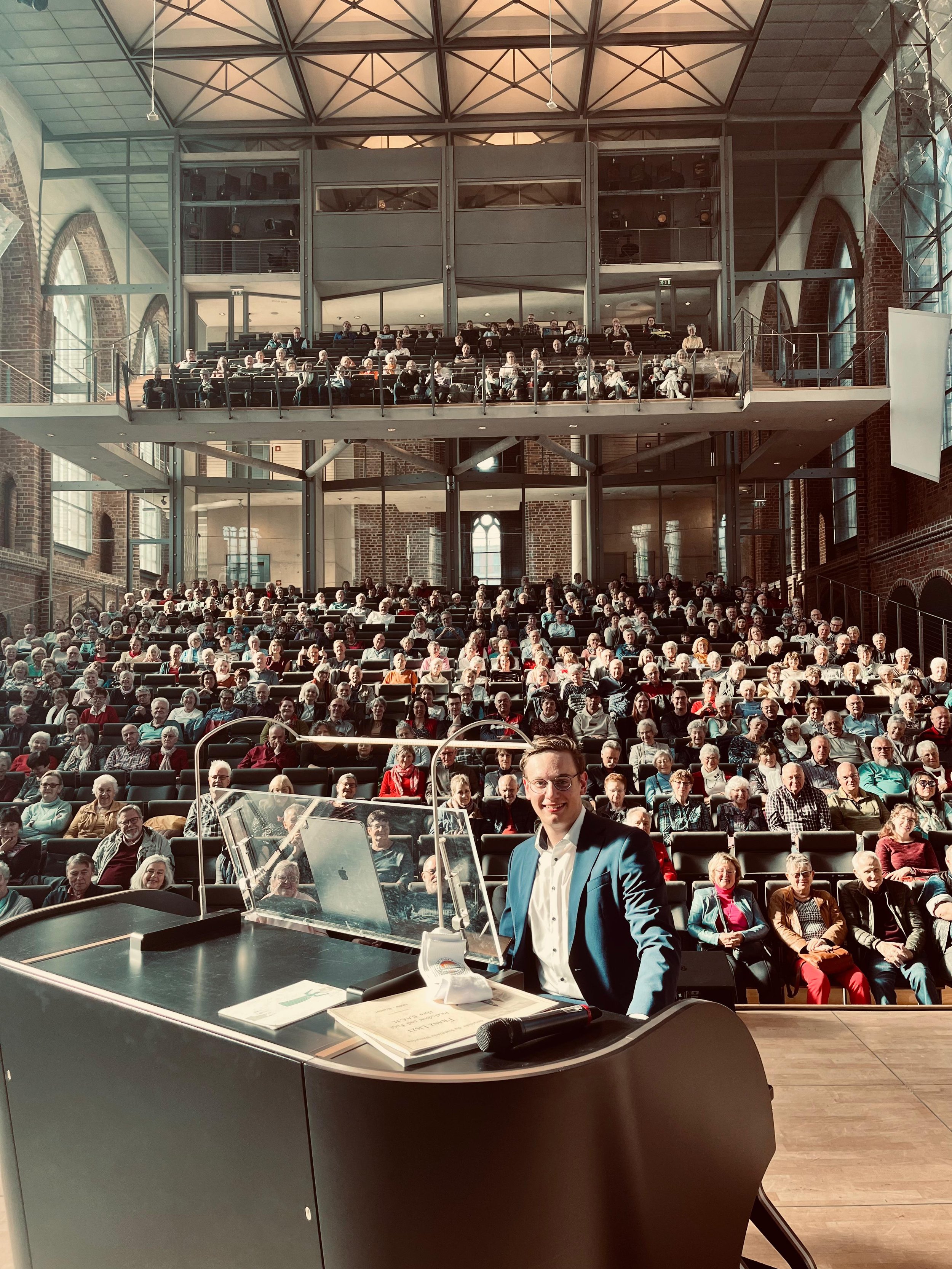 Ein Mann steht vor einem Podium in einem großen, modernen Veranstaltungssaal mit vielen Zuschauern. Der Saal hat hohe, große Fenster, die viel Tageslicht durchlassen, und eine hohe, gewölbte Decke.