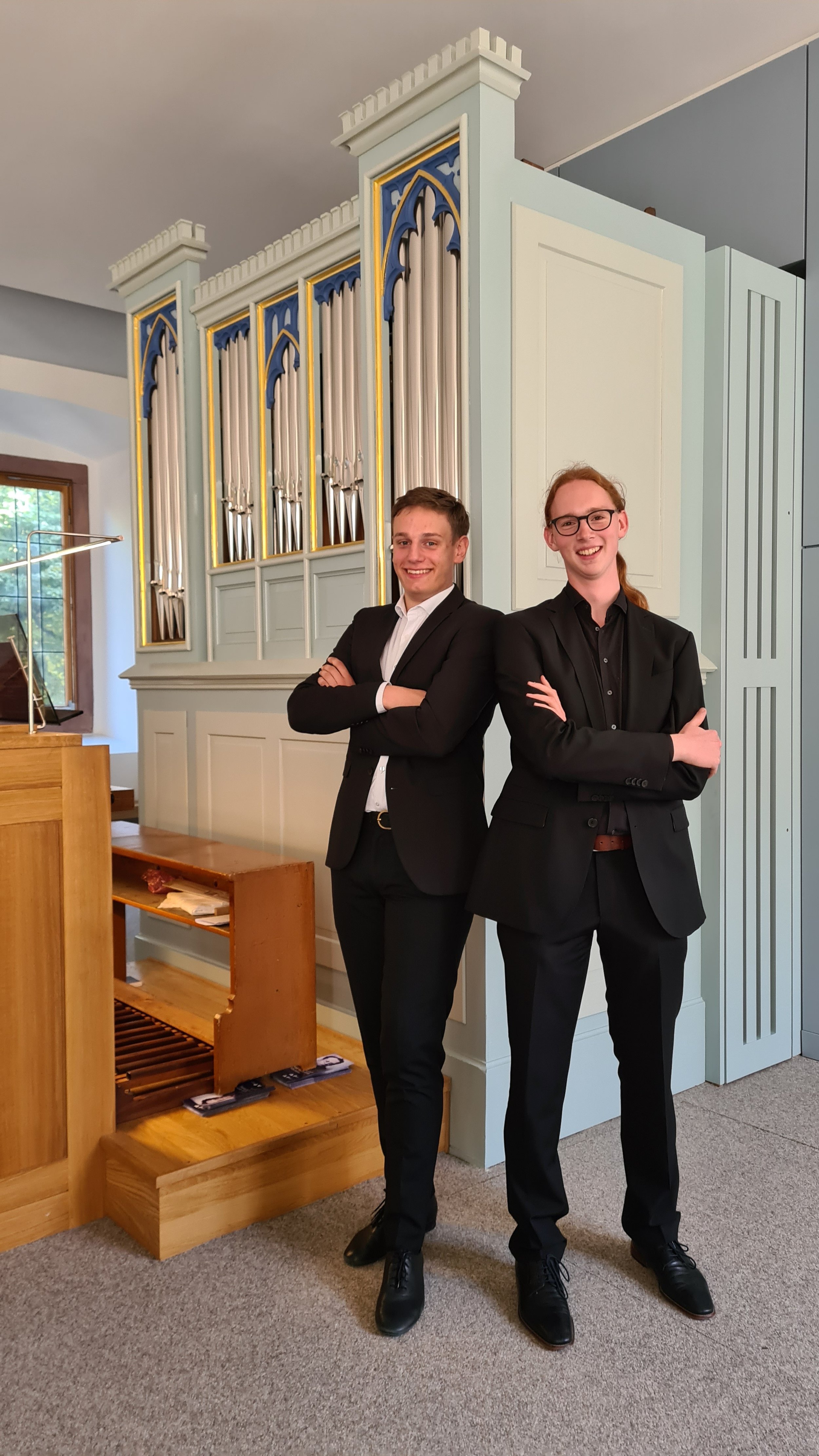 Zwei Jugendliche in schwarzen Anzügen stehen vor einer historischen Orgel in einem Raum, lächeln und haben die Arme verschränkt.