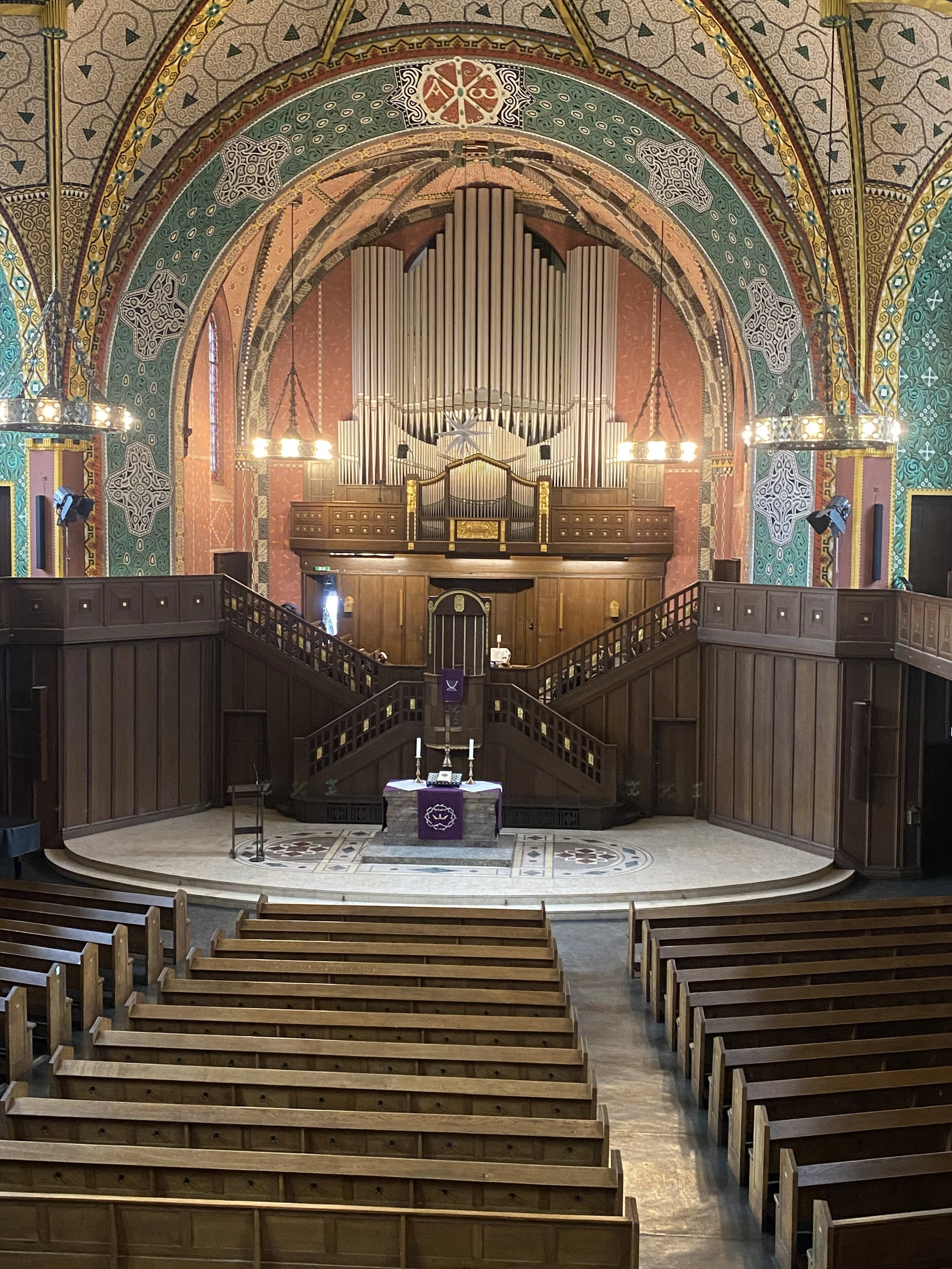 Innenansicht einer kirchlichen Kapelle mit Holzbänken, Altar, Treppen, einem großen Orgel und bunten Wand- und Deckengestaltungen.