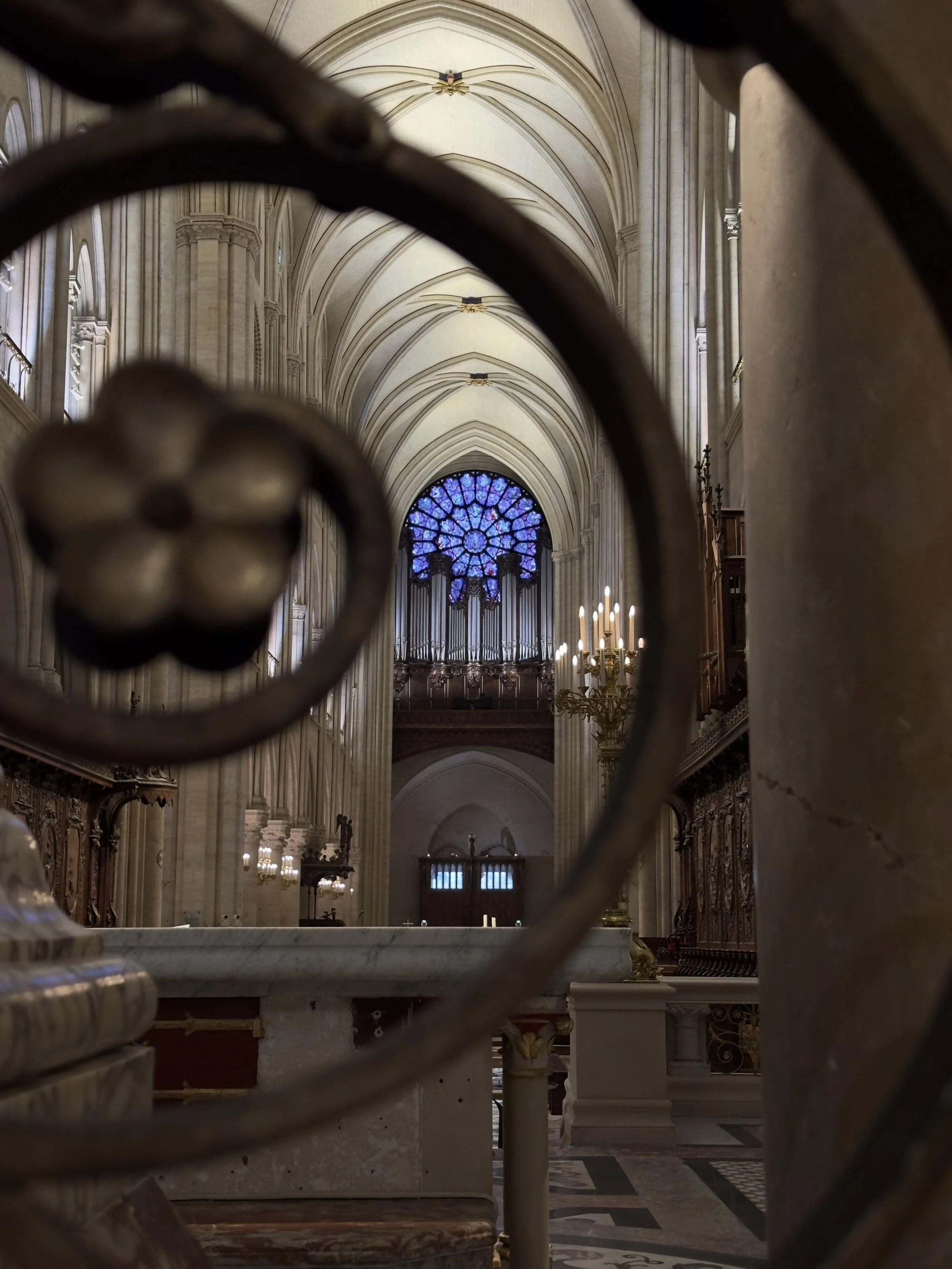 Blick durch eine kunstvoll verzierte Gitteröffnung auf den Innenraum einer gotischen Kathedrale, mit hoher, gewölbter Decke, einem bunten Rosettenfenster und Kerzenleuchtern.
