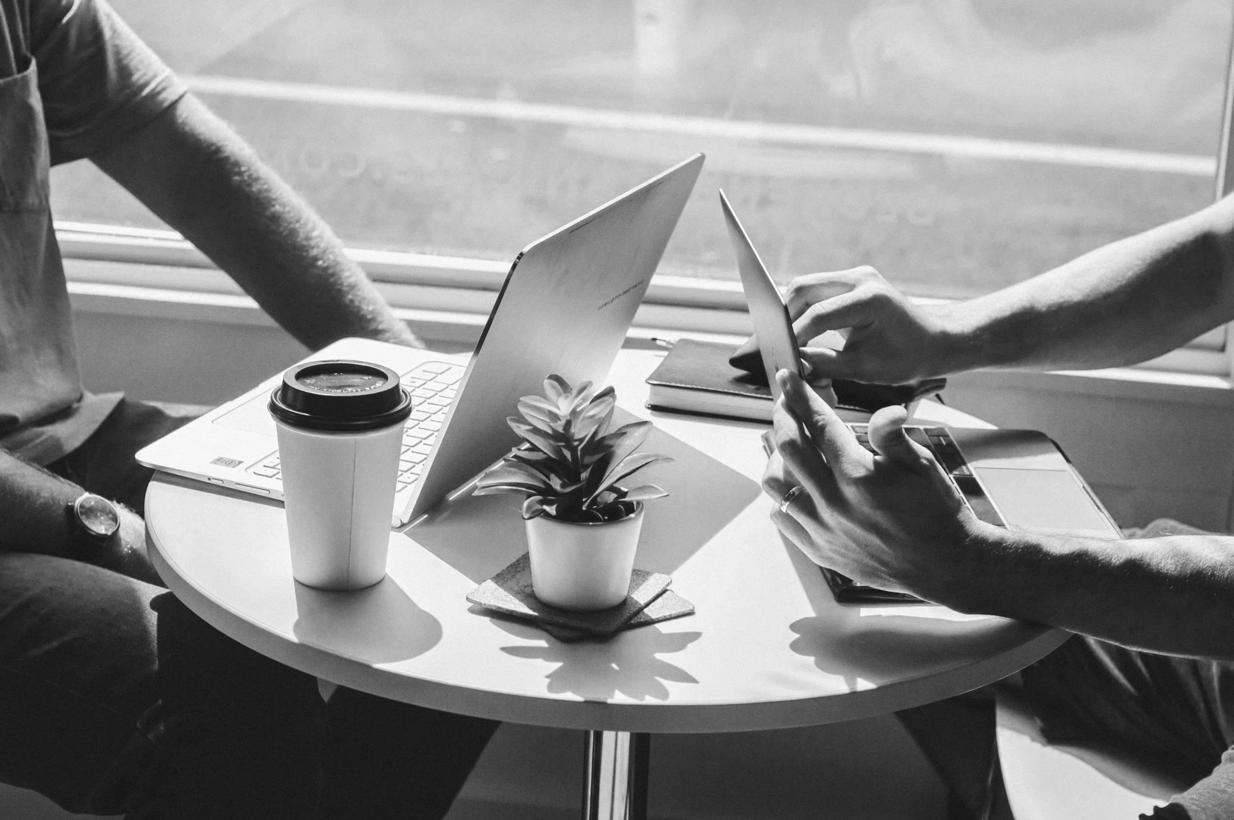 Two people sitting at a circular table near a window, working on a laptop and a tablet. The table has a potted plant, a coffee cup, and a closed notebook.