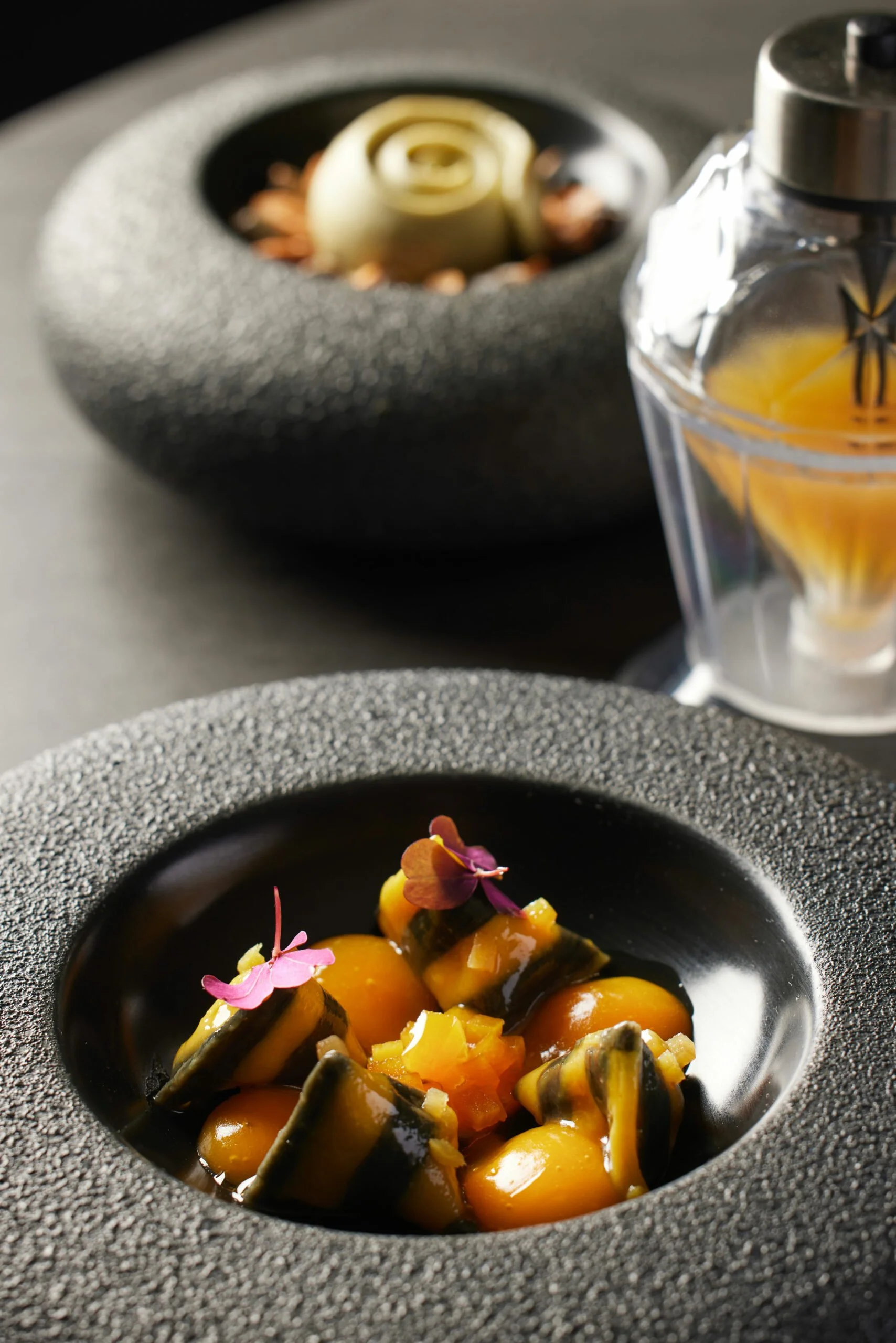 Assiette noire avec un plat de nourriture colorée garnie de fleurs comestibles. En arrière-plan, un autre plat ou décoration, et à droite, un flacon de parfum transparent avec un liquide jaune.