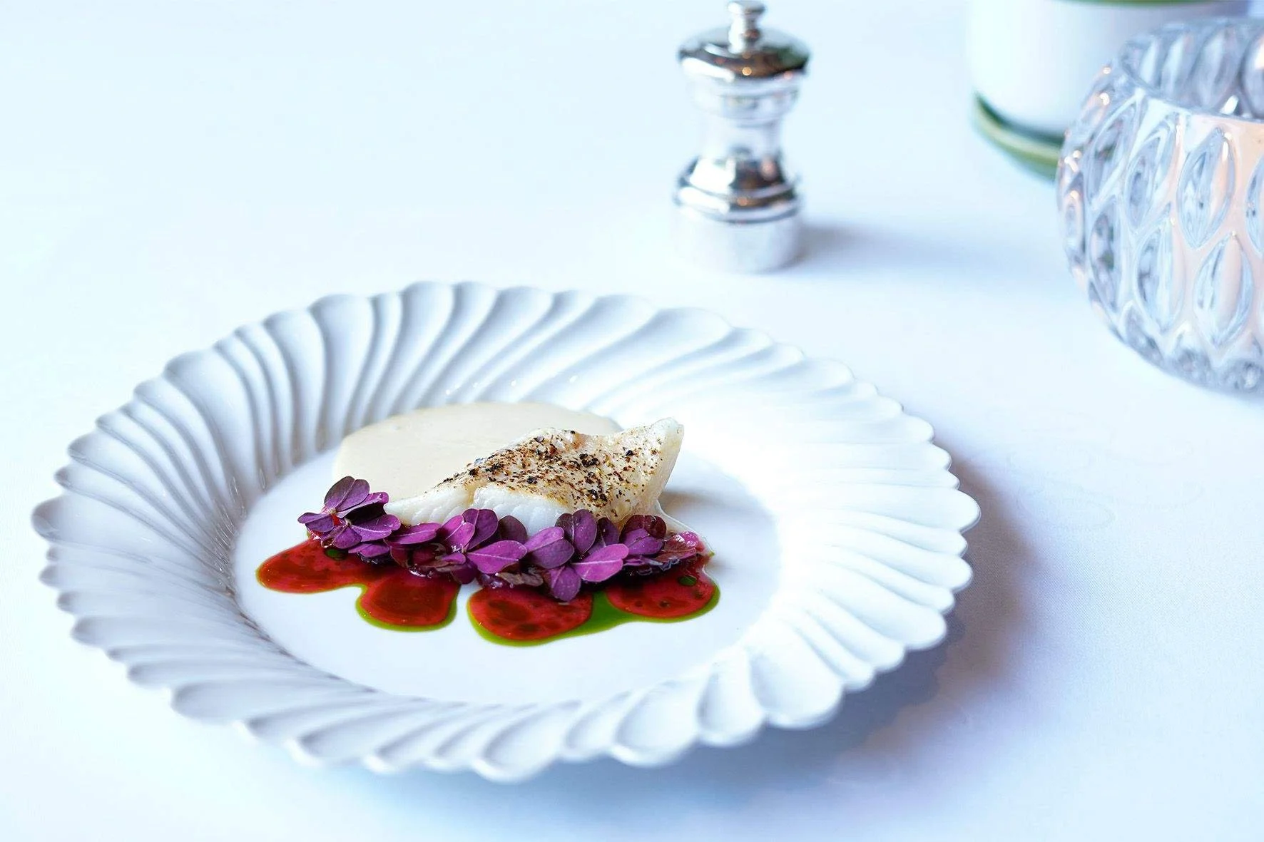 Assiette blanche avec un plat élégant comprenant un filet de poisson, des éclats de lavande violette, une sauce rouge, et une sauce verte, sur fond blanc avec un moulin à poivre en arrière-plan.