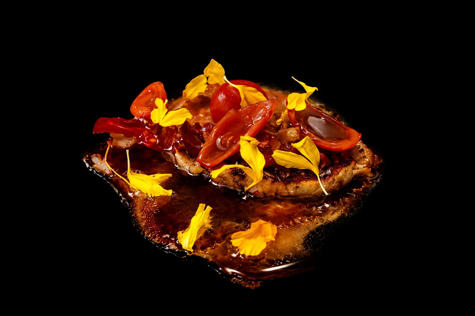 Une assiette de nourriture garnie de tomates cerises, de légumes et de fleurs comestibles, avec une sauce ou un jus au fond, sur un fond noir.