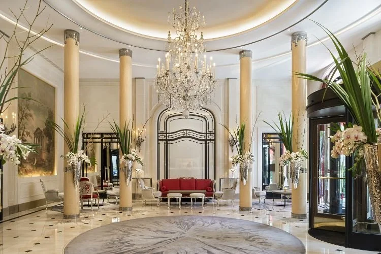 Intérieur élégant d'un lobby ou d'une salle de réception avec un grand chandelier, des plantes vertes, un canapé rouge au centre, et des colonnes en marbre, décorée dans un style classique et luxueux.