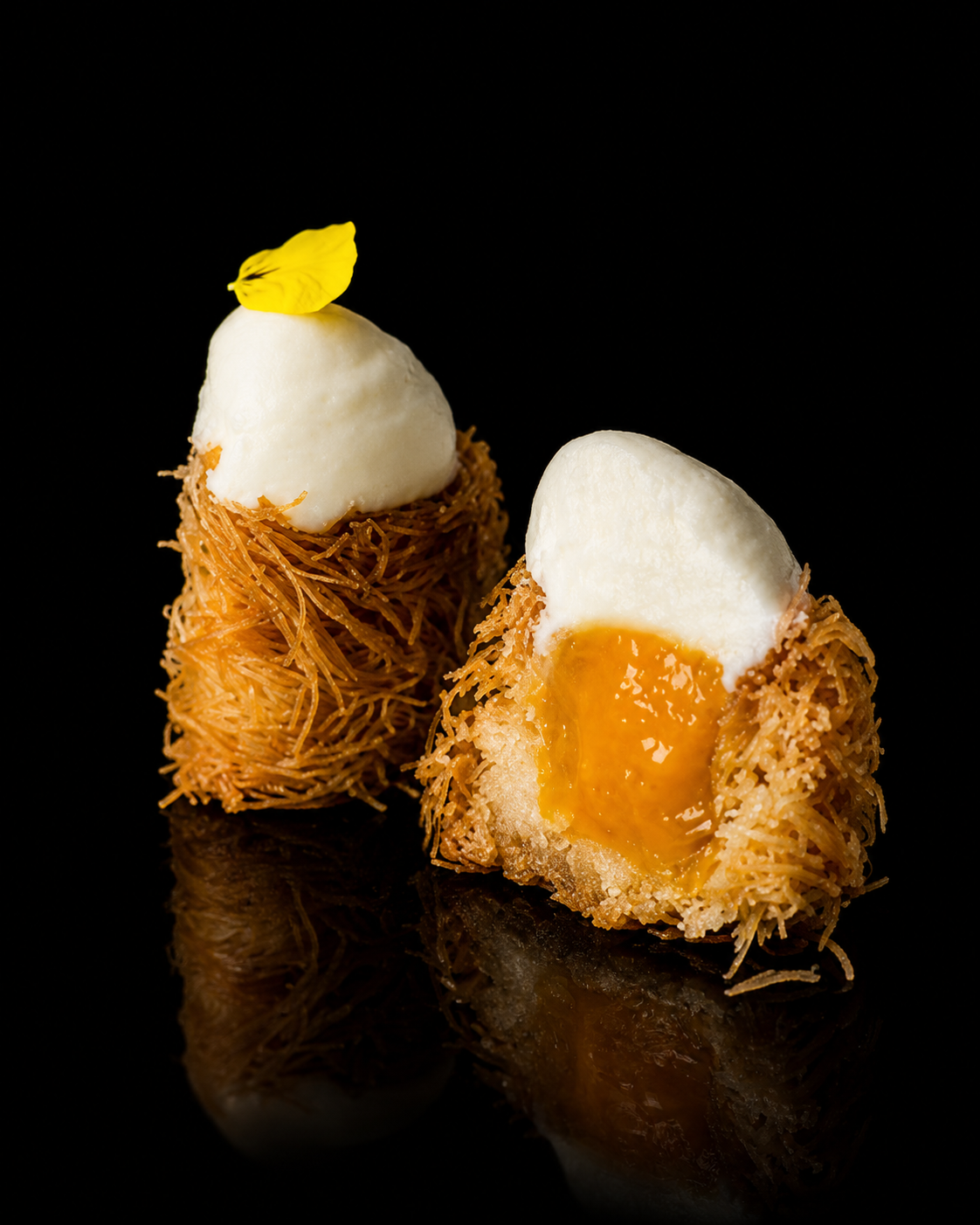 Deux profiteroles en nid de pâte croustillante, garnies de crème et de caramel, une décorée d'une fleur jaune sur le dessus, présentées sur un fond noir.