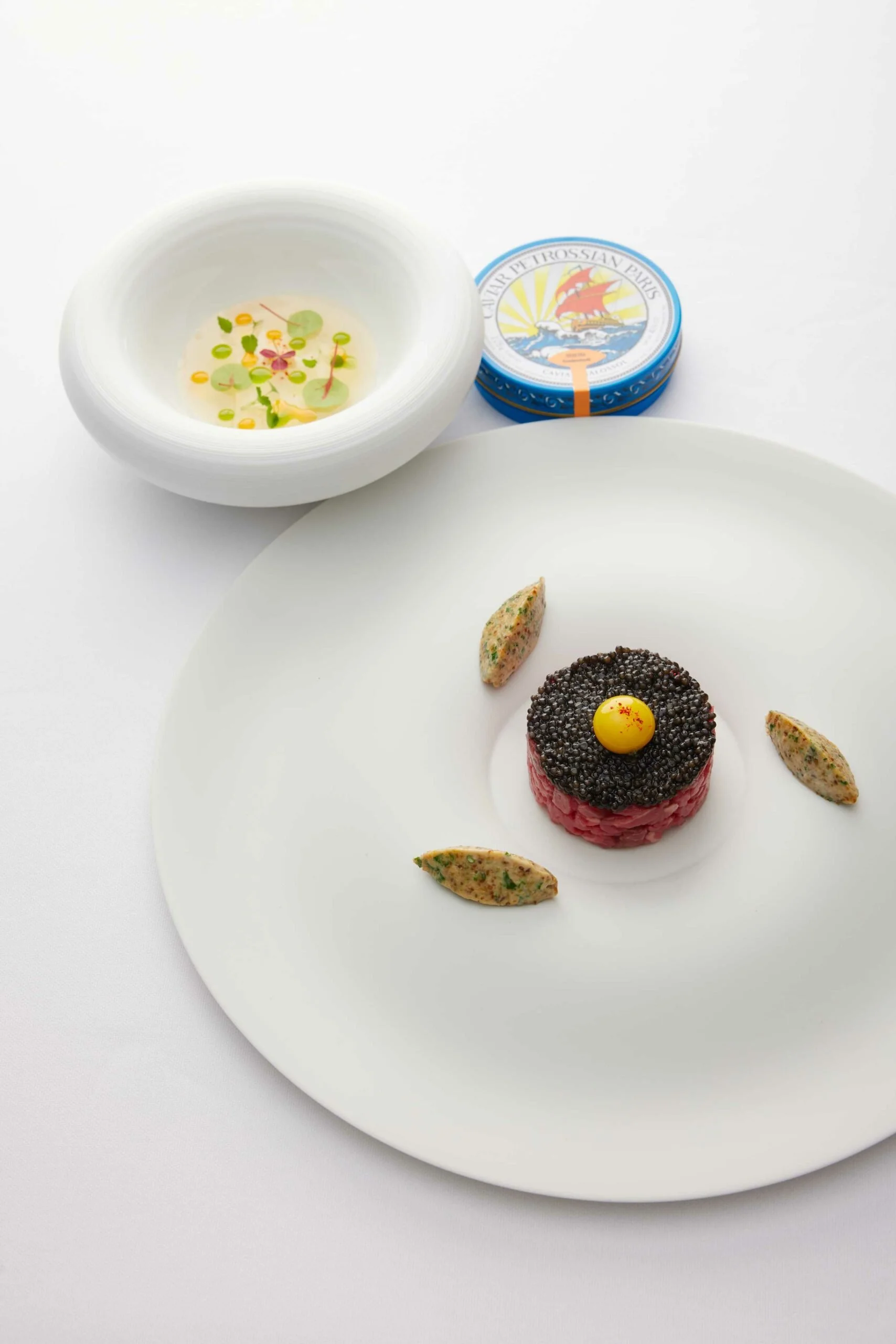 Assiette de tartare de viande garnie de caviar noir, œuf en dégradé de jaune, et petites quenelles, accompagnée d'une coupe de soupe claire avec des herbes et d'une boîte de tartare.