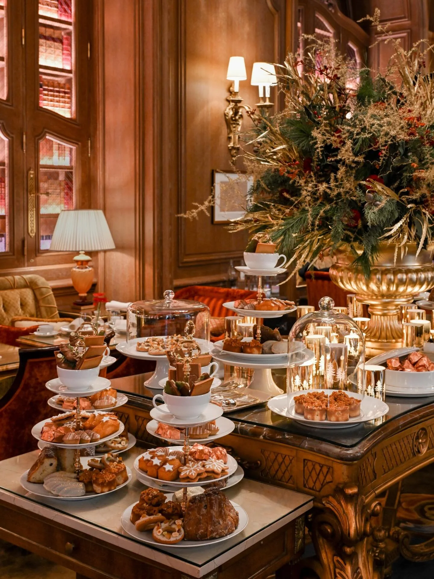 Table décorée pour un thé de l'après-midi avec des pâtisseries, biscuits et plateaux, dans une pièce élégante avec bois sombre et éclairage chaud.
