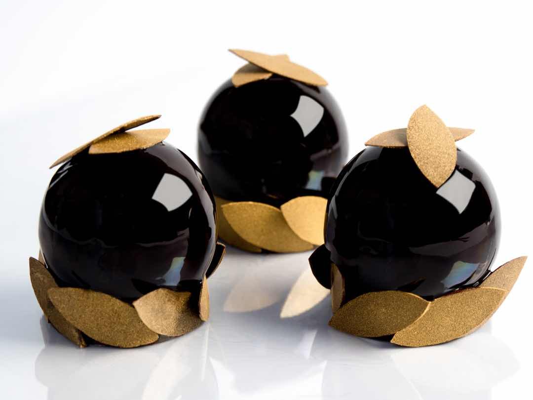 Trois desserts sphériques noirs avec des décorations dorées en forme de feuilles enroulées autour.