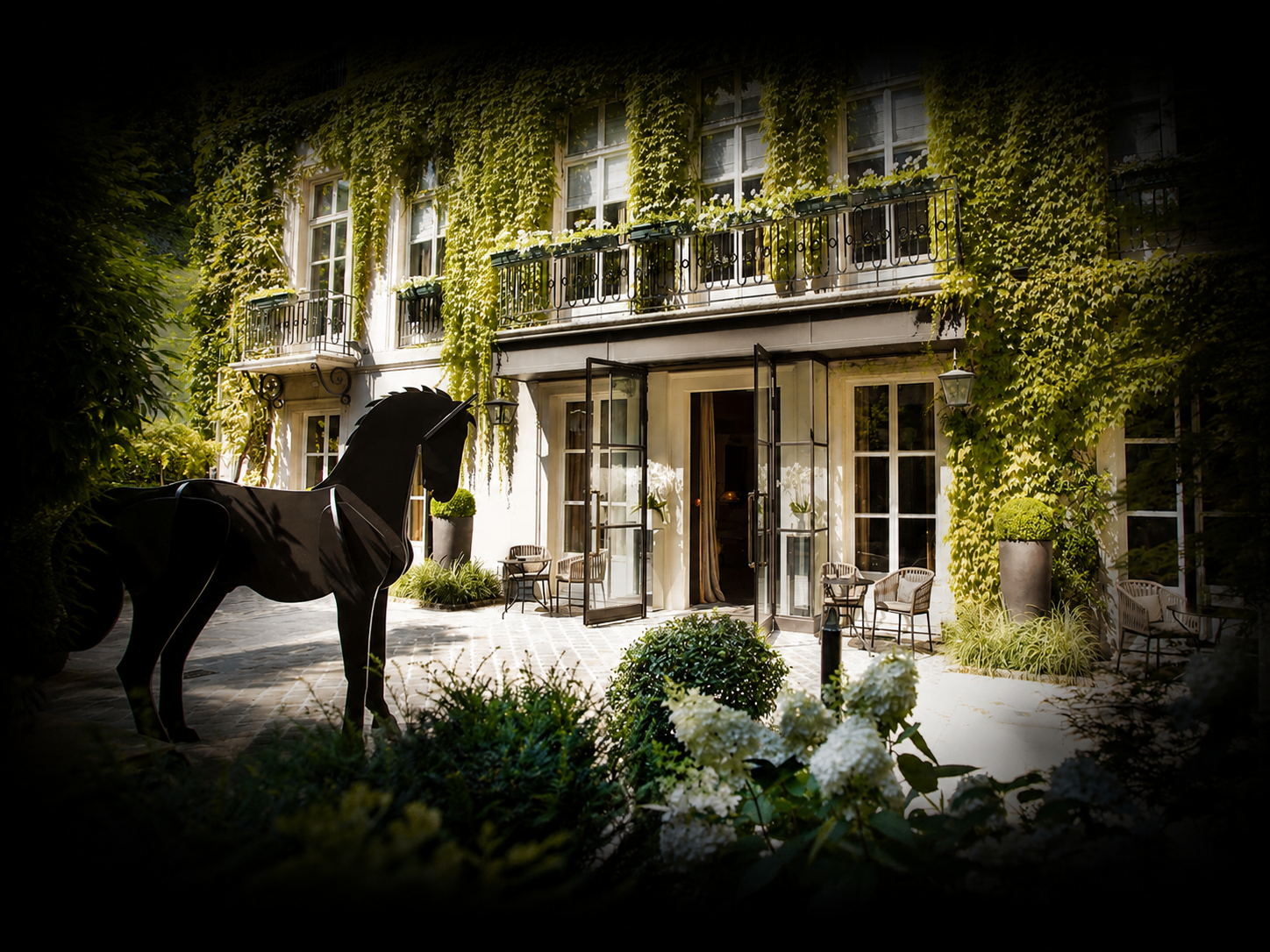 Façade d'un bâtiment en pierre avec des balcons en fer forgé, recouvert de vignes vertes. Un cheval en silhouette noire se dresse devant l'entrée principale, entouré de plantes et de fleurs blanches, avec des chaises à l'extérieur.