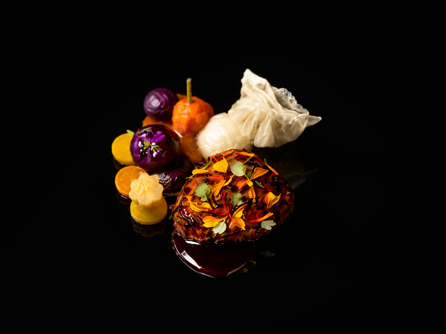 Un plat gourmet avec différents éléments colorés, dont une pièce principale recouverte de fleurs et d'herbes, accompagnée de légumes en petits morceaux et de raviolis sur un fond noir.
