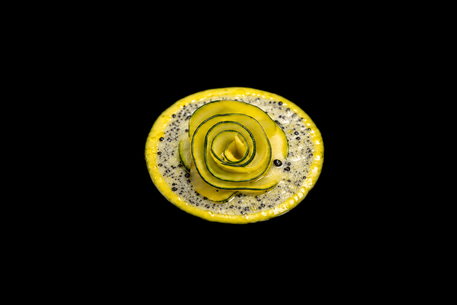 Fleur décorative faite de rondelles de concombre enroulées en forme de rose, posée sur une tranche de citron avec des graines de lin et de chia, sur un fond noir.