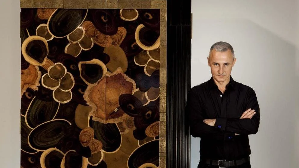 Un homme debout avec les bras croisés à côté d'une œuvre d'art abstraite composée de cercles et de formes organiques dans des tons marron, beige, et noir.