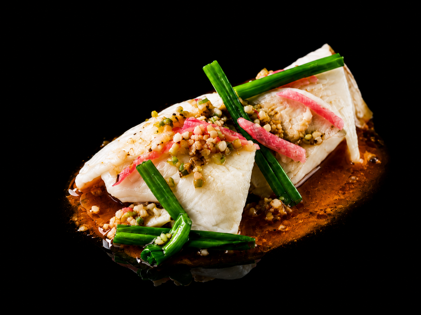 Filet de poisson blanc garni de graines et de fines légumes, servi avec une sauce sombre, présenté sur un fond noir